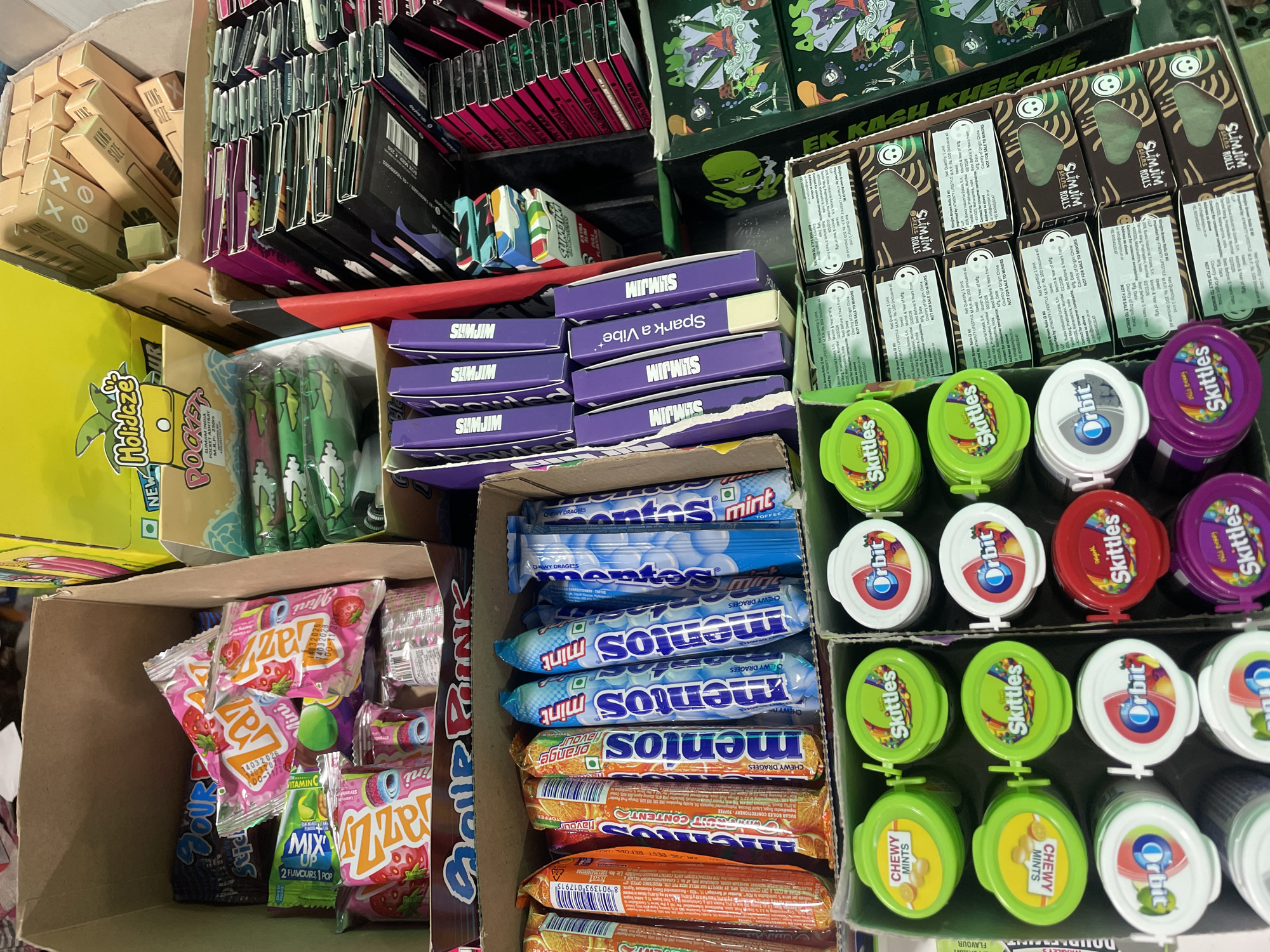 The image shows a retail display of various confectionery items, including candies and gum, arranged in cardboard boxes and on shelves. The items are predominantly packaged in colorful wrappers and containers. Several brands are visible, such as Mentos, Orbit, Skittles, and others in colorful packaging. Some of the items appear to be mints, while others are fruity candies. The overall scene suggests a store or market stall. The image is taken from a slightly elevated angle, looking down into the display. The lighting suggests an indoor environment. There are no people visible in the image.