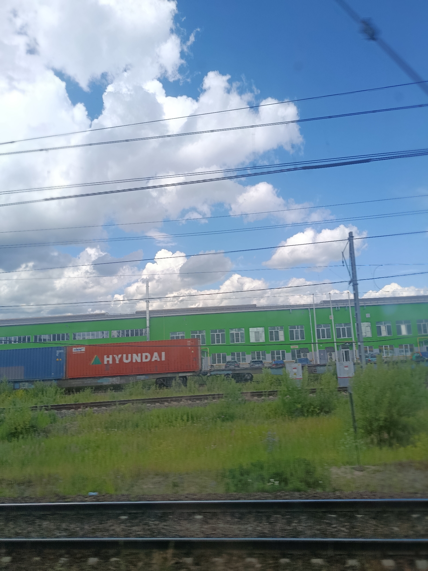 The image shows a bright, cloudy blue sky above a long, green industrial building. In front of the building, there are railway tracks with several cargo containers, including a prominent orange container with "HYUNDAI" written on it in white lettering and a blue container next to it. The building has numerous windows and appears to be a commercial or industrial facility. The foreground shows more railway tracks and overgrown green grass. The overall scene suggests a view from a moving vehicle, likely a train, passing by an industrial area on a sunny day. This location is identified as Bulagovo-Life, Russia.