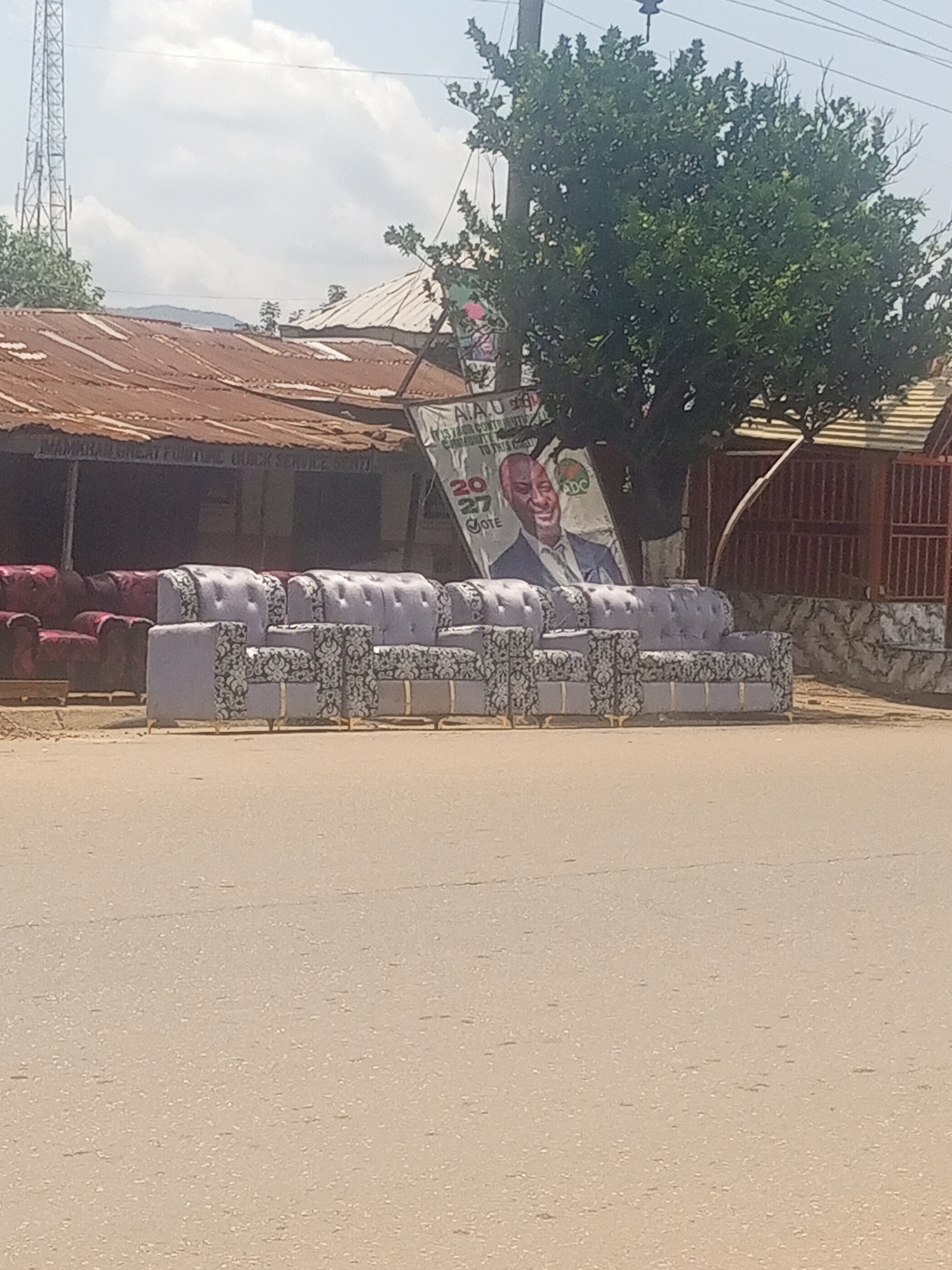 The image shows a street scene in Serti, Nigeria, with a row of upholstered sofas displayed for sale outside a shop. The shop has a rusty corrugated metal roof. Behind the sofas, a large poster featuring a smiling man in a suit is visible, along with some political slogans. A tall telecommunications tower stands in the background to the left, and a lush green tree dominates the right side of the frame. The sky is partly cloudy. The overall impression is a commercial area with outdoor furniture display.
