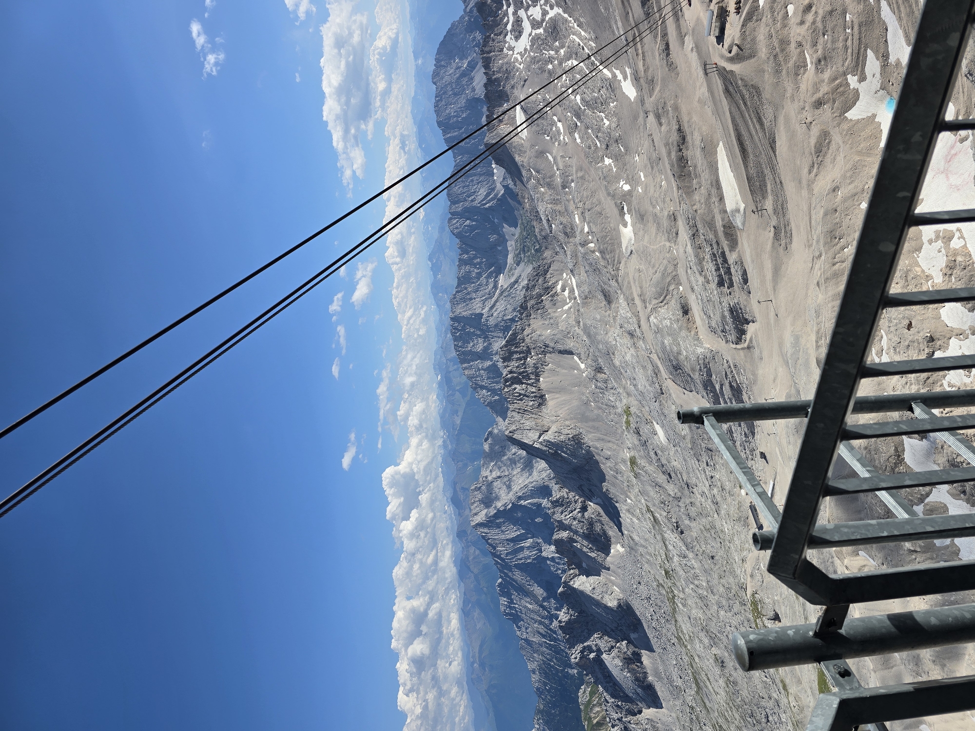 The image depicts a panoramic view of a mountainous landscape, likely taken from a high vantage point, possibly a cable car or viewing platform. In the foreground, a metal railing and support posts create a barrier, suggesting a designated viewing area. Three dark cables extend diagonally across the upper portion of the frame, indicating the presence of a cableway system.
The mid-ground and background are dominated by rugged, rocky mountain ranges, some of which are covered in patches of snow. The terrain appears arid and stony, with evidence of hiking trails or ski slopes etched into the landscape. A small building or facility is visible on the right side of the mid-ground, near a ski lift. The sky is a vibrant blue with scattered white clouds.
The scene is characteristic of the Zugspitze region near Grainau, Germany, a popular destination for mountain tourism known for its cable car access and scenic vistas. No people are visible in the image.