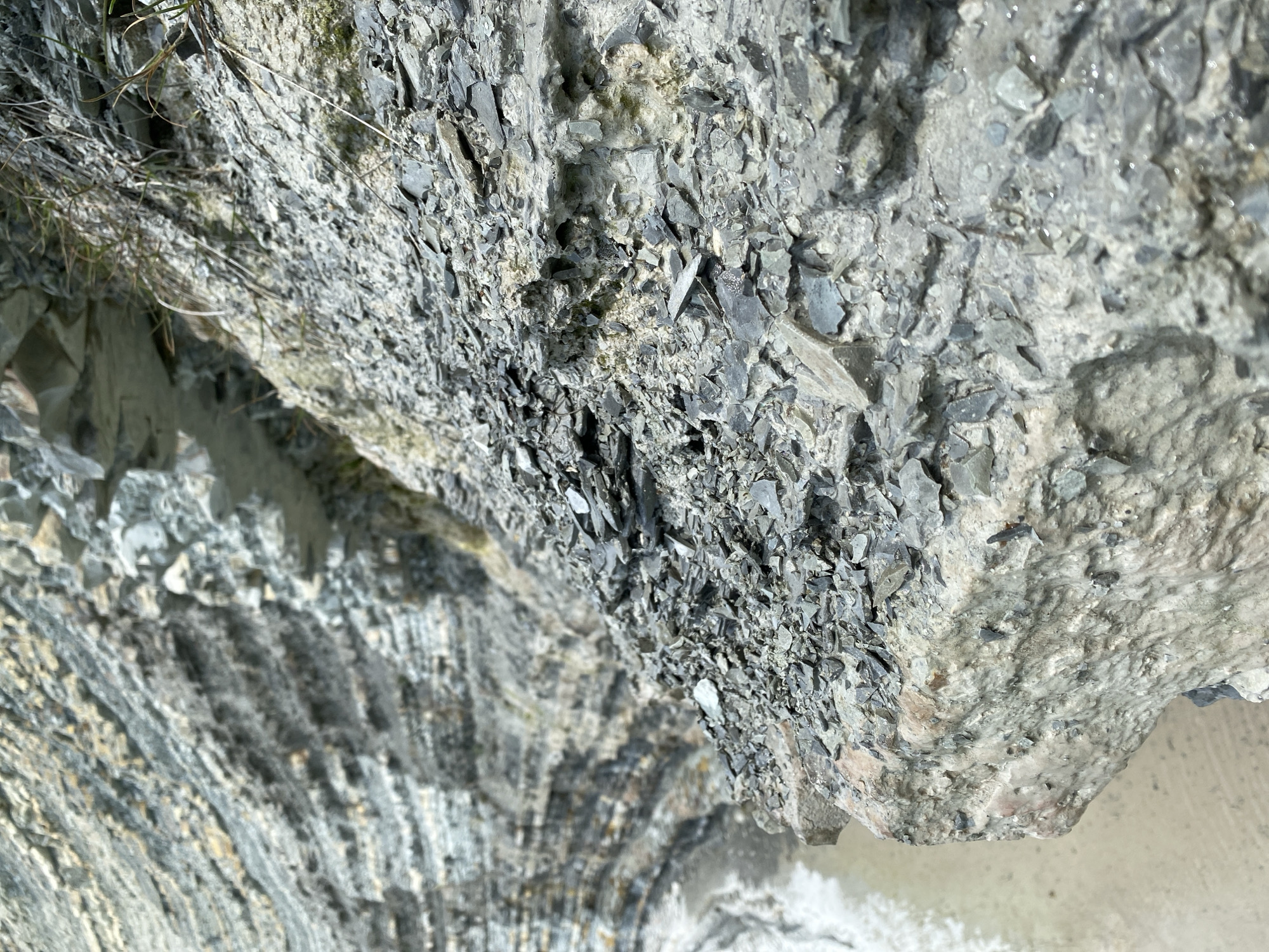 The image presents a detailed, angled close-up of a weathered rock formation, likely part of a cliff or riverbank. The dominant features are the varying textures and compositions of the rock, illuminated by bright daylight.
The upper and central portions of the image are characterized by a dense accumulation of loose, angular, dark grey to blackish-grey rock fragments, resembling shattered shale or slate. These pieces vary in size, creating a jagged, uneven surface. Interspersed within these fragments are patches of lighter, pale grey to off-white rock, which appears more solid but also heavily eroded and crumbly in places. Some of the rock surfaces, particularly among the loose fragments, exhibit a glistening sheen, suggesting dampness or recent exposure to water.
Along the upper-left edge, sparse, dry strands of grass or roots cling to the rock, indicating minimal vegetation.
Towards the lower-left, the rock formation transitions into a section displaying distinct, wavy, horizontal layers or striations. These layers alternate in shades of dark grey, lighter grey, and beige, showcasing geological stratification. This part of the rock appears more consolidated and less fragmented than the upper sections.
The very bottom-right corner of the image reveals a lighter-colored, flatter area, possibly sand, silt, or shallow water, suggesting the base of the rock face. There are no people, animals, or artificial objects visible, and no discernible text. The scene captures a natural, static geological landscape.
