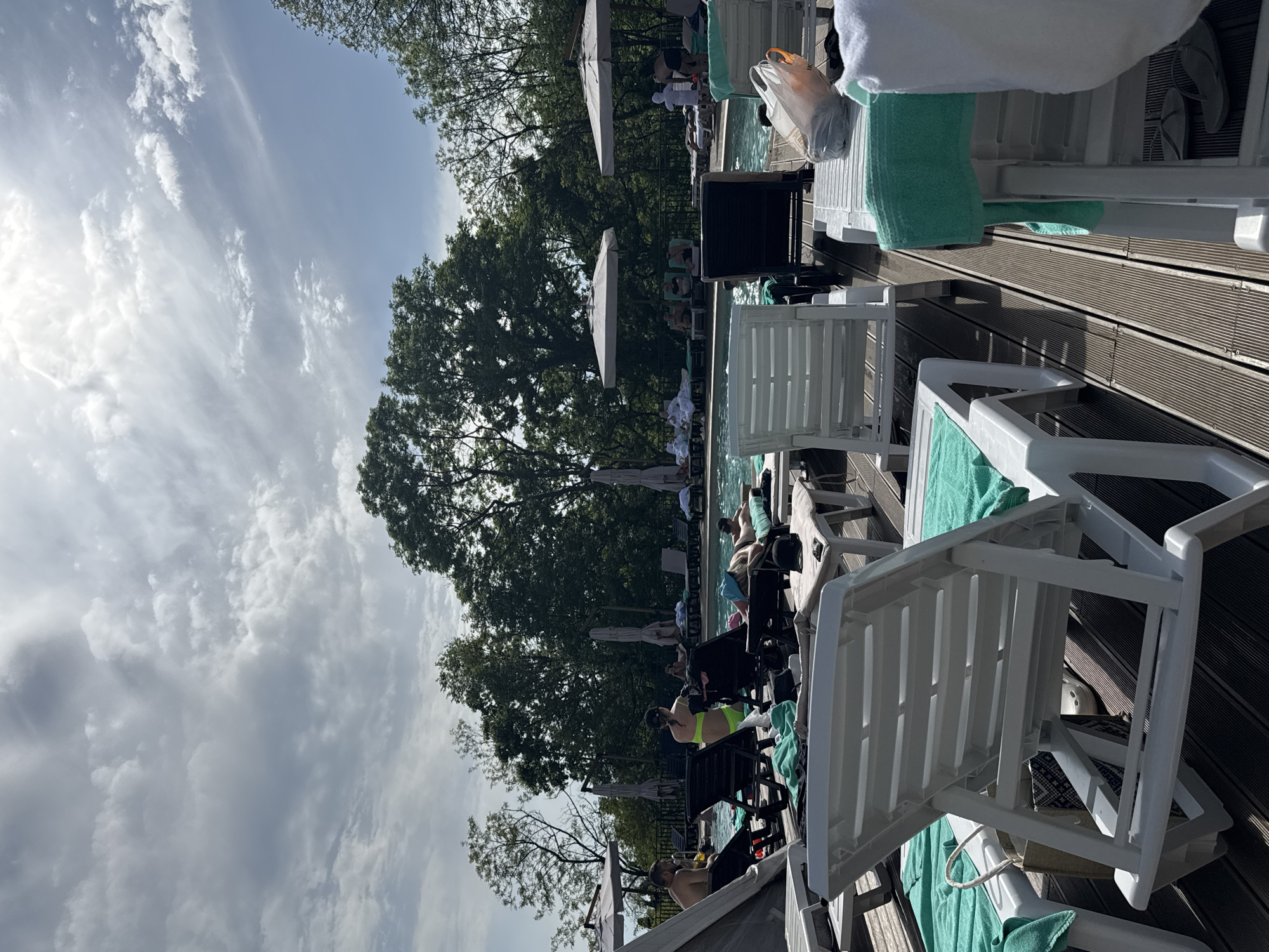 The scene depicts a swimming pool area in Sosnovy, Russia, populated with individuals relaxing on lounge chairs adjacent to the water. Lounge chairs, both white and black, are aligned on a wooden deck surface. Some chairs have towels draped over them, while others are occupied by people in swimwear, some of whom are interacting. Umbrellas provide shade. Trees are visible in the background, partially obscuring a cloudy sky. The orientation is vertical.