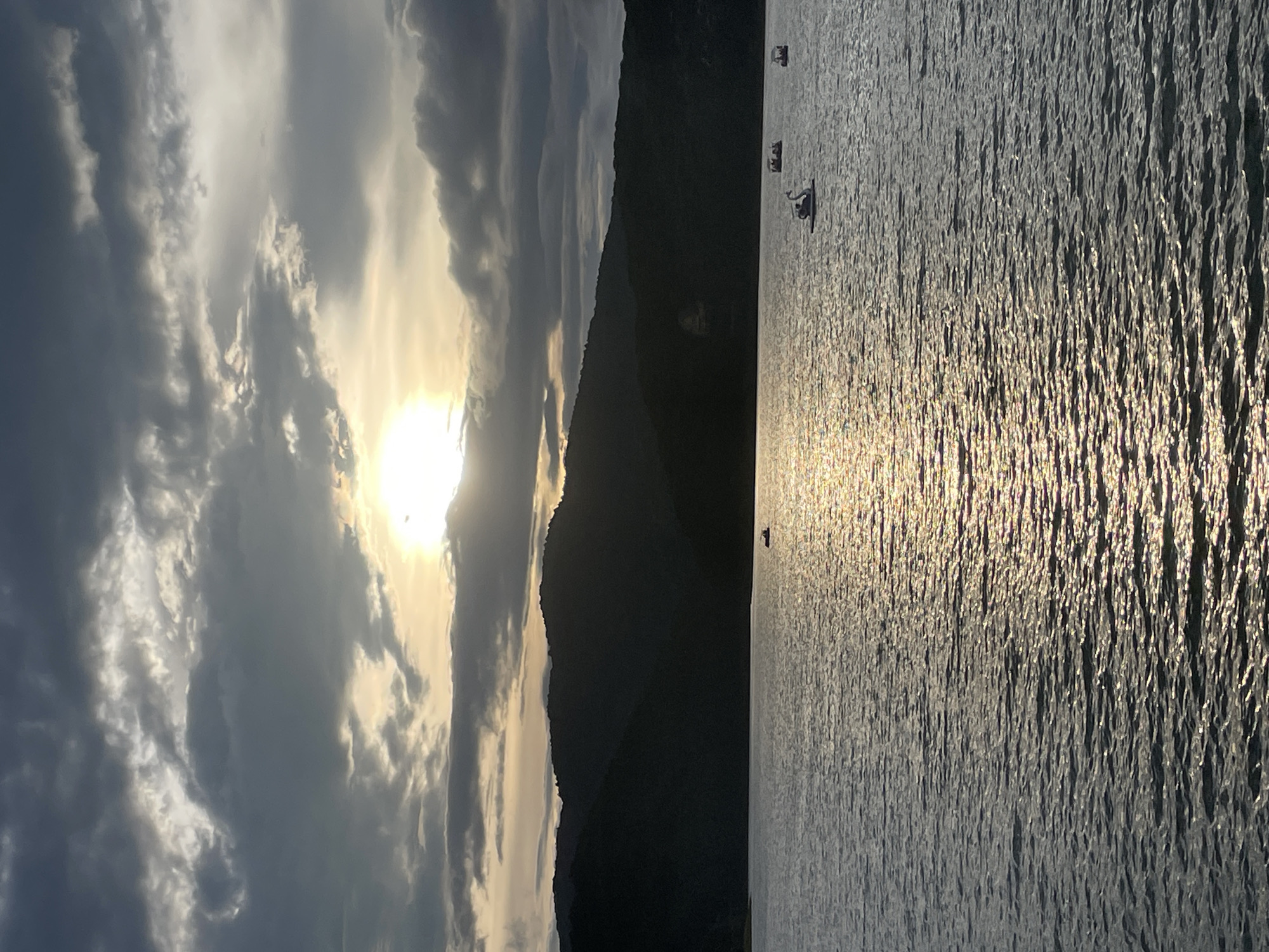 This image depicts Xuan Huong Lake in Da Lat, Vietnam, at dusk. The sun, partially obscured by dramatic, dark clouds, casts a shimmering golden reflection on the rippling water. Silhouetted against the sky are the rolling hills that surround the lake, their dark forms creating a stark contrast with the illuminated clouds. Scattered across the water are several pedal boats, including one shaped like a swan, each carrying a small group of people. The overall atmosphere is serene and picturesque, characteristic of Da Lat's natural beauty.