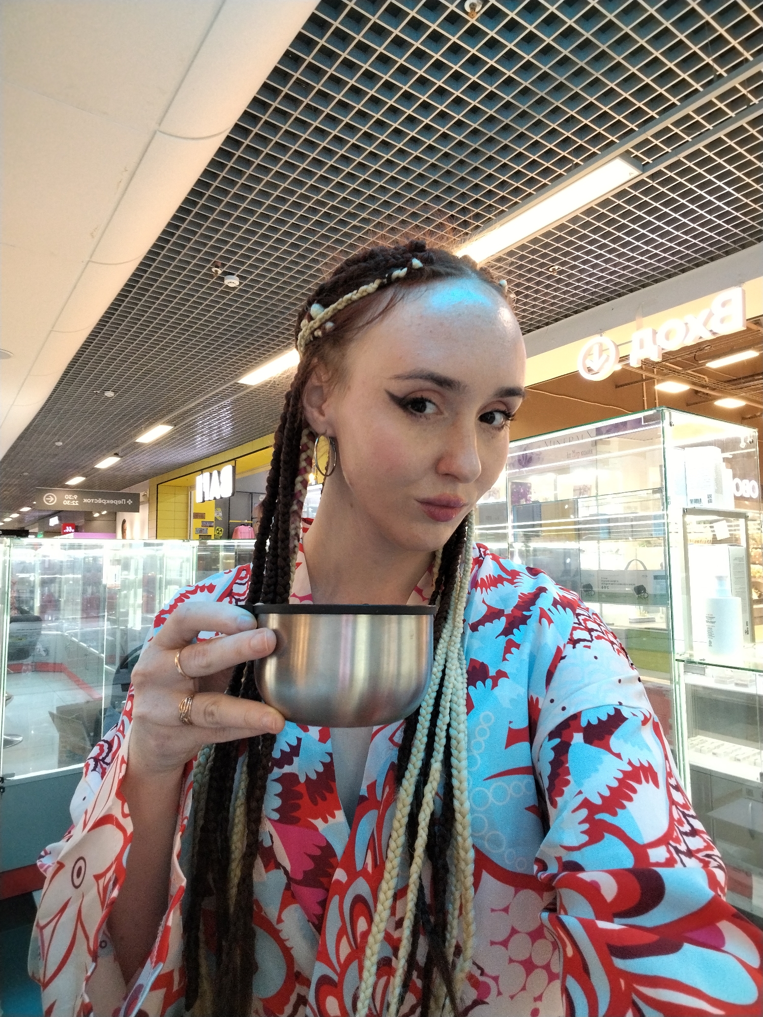 The image shows a woman holding a silver cup. She is wearing a colorful floral-patterned garment and has long braids, some blonde and some brown. She wears gold hoop earrings, a decorative band in her hair, and rings on her fingers. She is looking at the camera with a slight pout.
The background suggests an indoor public space, possibly a shopping center, with a grid-patterned ceiling. Display cases with products are visible behind her. There are also signs in the background, one which appears to say "Дона."
The image was taken in the city of Izhevsk, Russia.