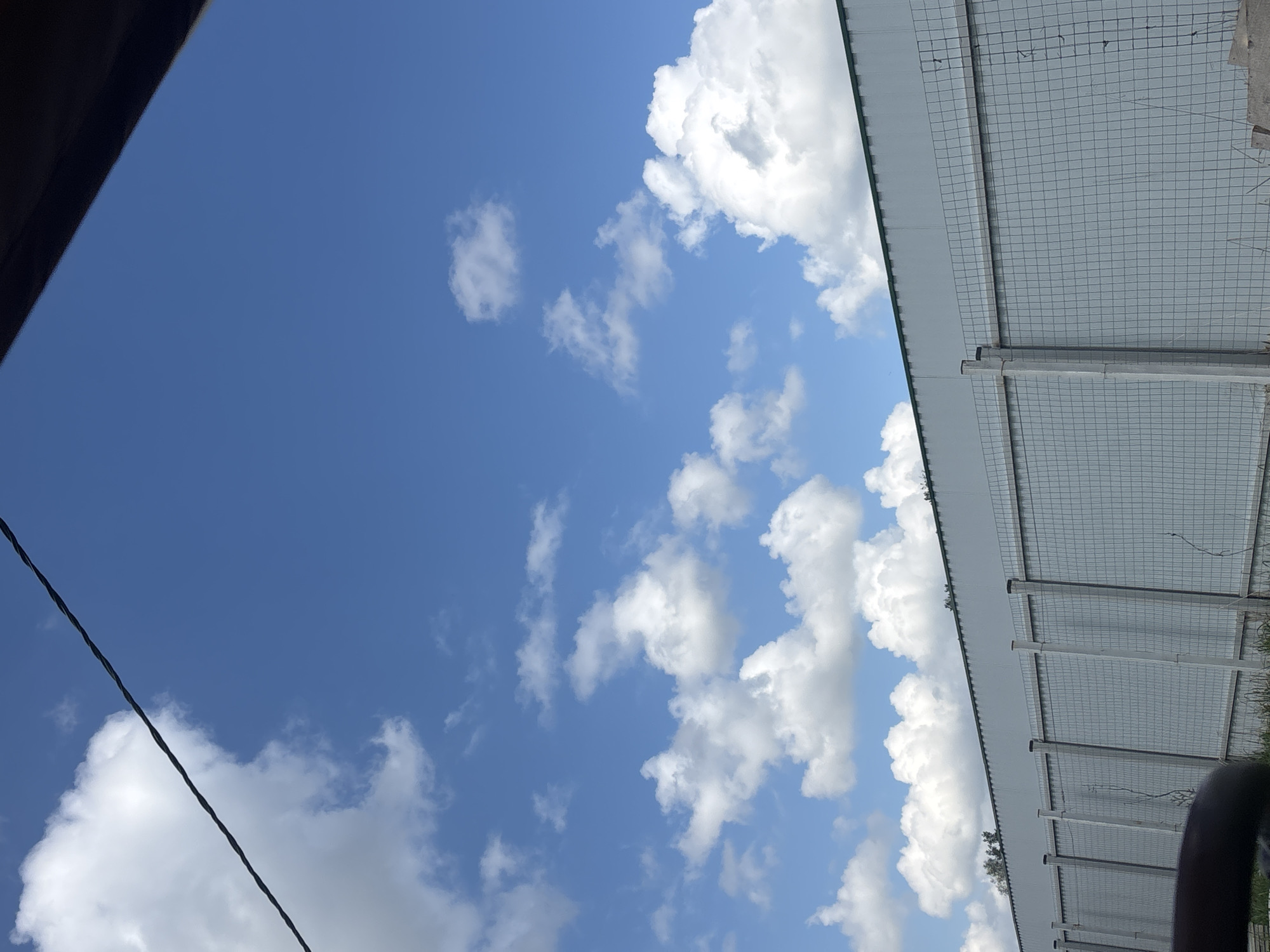 The image shows a view looking up at a bright blue sky with scattered white clouds. A dark object is visible in the upper left corner, partially obscuring the sky. In the upper right portion of the image, there is a structure with a gray, metallic roof and a wire mesh side. An electrical wire cuts across the lower left quadrant of the sky. The location is presumably Раздолье, Russia.