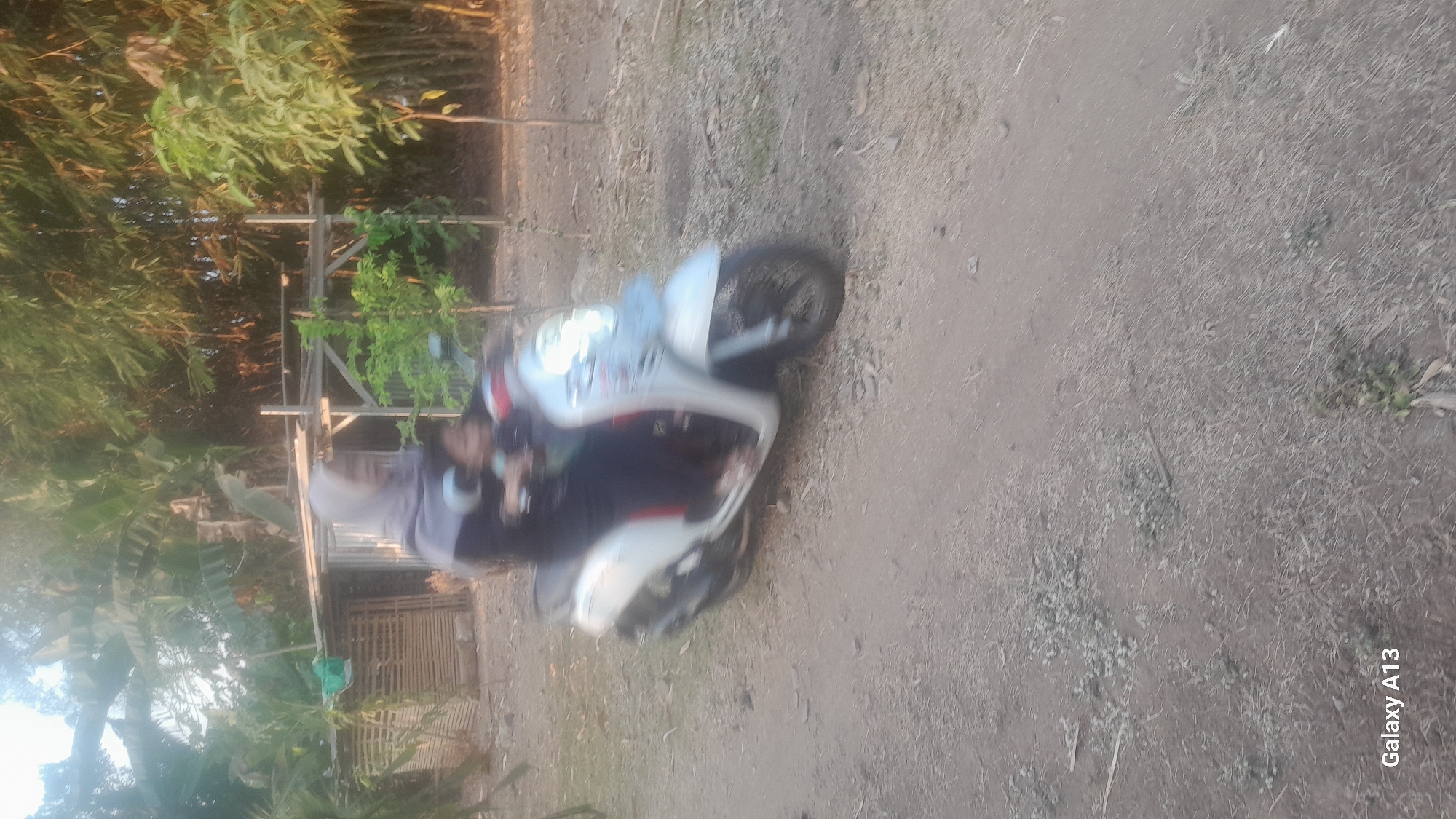 A woman wearing a grey hijab and a dark outfit rides a white scooter. She is carrying a child on her lap. The scooter is in motion, indicated by motion blur. The background consists of vegetation, including banana trees and bamboo, as well as a rustic structure. The ground is unpaved and appears to be dirt with small stones and dry grass. The scene is outdoors, likely in a rural or semi-rural area. The image is also watermarked with "Galaxy A13" in the lower left corner.