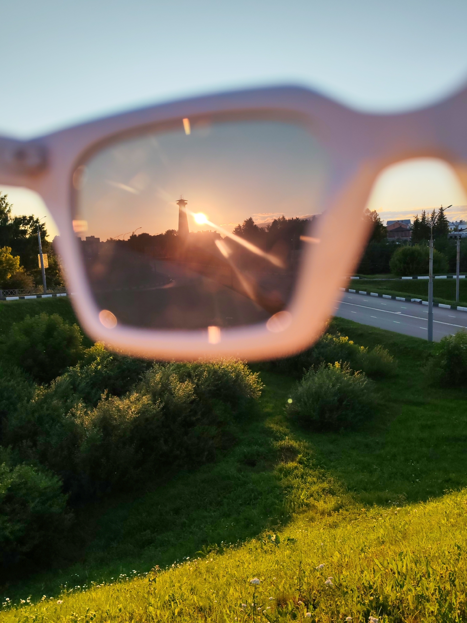 This is a vertically oriented, medium shot image taken from the perspective of looking through a pair of light pink framed sunglasses at sunset. The foreground is out of focus, with the sunglasses frames creating a bright, blurred border around the central subject. The lenses of the sunglasses are in focus and reflect the scene.
The reflected scene depicts a sunset with the sun positioned slightly to the left of the center, casting a warm orange glow and lens flares across the frame. A tall, slender tower structure with a platform at the top is visible in the distance, silhouetted against the bright sky. The landscape within the reflection includes rolling hills covered in lush green trees and bushes, with a winding road visible in the middle ground to the right, bordered by a white and black striped curb and grass. In the far background, rooftops of buildings and more trees can be seen against the hazy horizon. The sky is a gradient of pale blue at the top, transitioning to soft orange and pink hues near the horizon.
The area depicted is the city of Bol'shie Mkhi, Russia. The environment suggests an outdoor setting with a mix of urban and natural elements, characterized by grassy slopes, dense vegetation, and infrastructure like roads and streetlights. There are no people or animals visible in the image. The overall mood is serene and atmospheric, with the sunset lighting enhancing the natural beauty of the landscape.