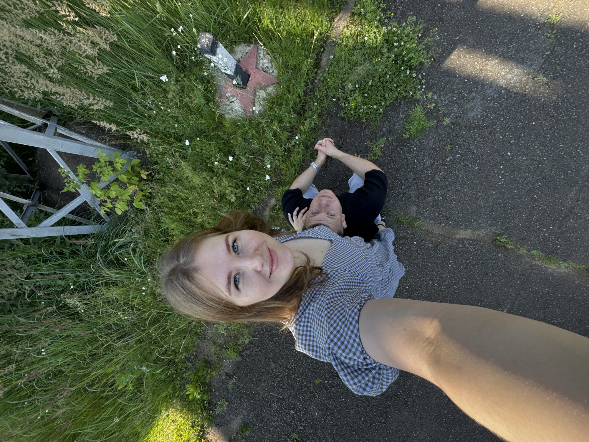 The image shows a high-angle selfie taken outdoors. In the foreground, a young woman with light brown hair and blue eyes is smiling and looking up at the camera. She is wearing a grey and white checkered dress. To her right and slightly behind her, a young man with short brown hair and blue eyes is also looking up at the camera. He is wearing a black t-shirt and grey shorts. They are standing on a dark asphalt path next to a grassy area with tall green grass and some small white flowers. In the background, there is a metal structure, possibly part of a power line tower, and a red and black star-shaped marker on a concrete base. The lighting suggests it is daytime with sunlight casting shadows. This scene is consistent with the area around the Perm-Prikamskaya substation on the banks of the Kama River in Perm, Russia, which is known for its industrial infrastructure and surrounding natural landscape.