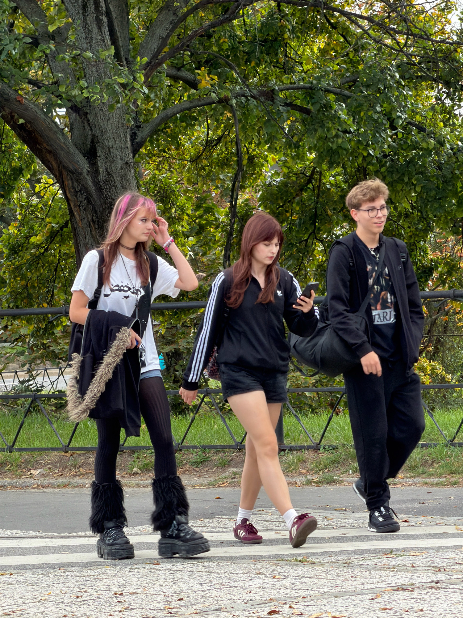 Three young people are walking across a pedestrian crossing in front of a park. The person on the left is a young woman with pink-streaked hair, wearing a white t-shirt, black fuzzy leg warmers over black tights, and large platform boots. She is carrying a black jacket with a furry trim over her left arm and a backpack. The person in the middle is a young woman with long, dark hair, wearing a black Adidas track jacket, black shorts, and burgundy sneakers. She is looking at her phone. The person on the right is a young man with short, light brown hair and glasses, wearing a black jacket over a t-shirt and black pants. He is carrying a black duffel bag over his shoulder. The background consists of trees and foliage, suggesting a park or green space. The pedestrian crossing is marked with white stripes on asphalt. The overall impression is of a casual outdoor scene.