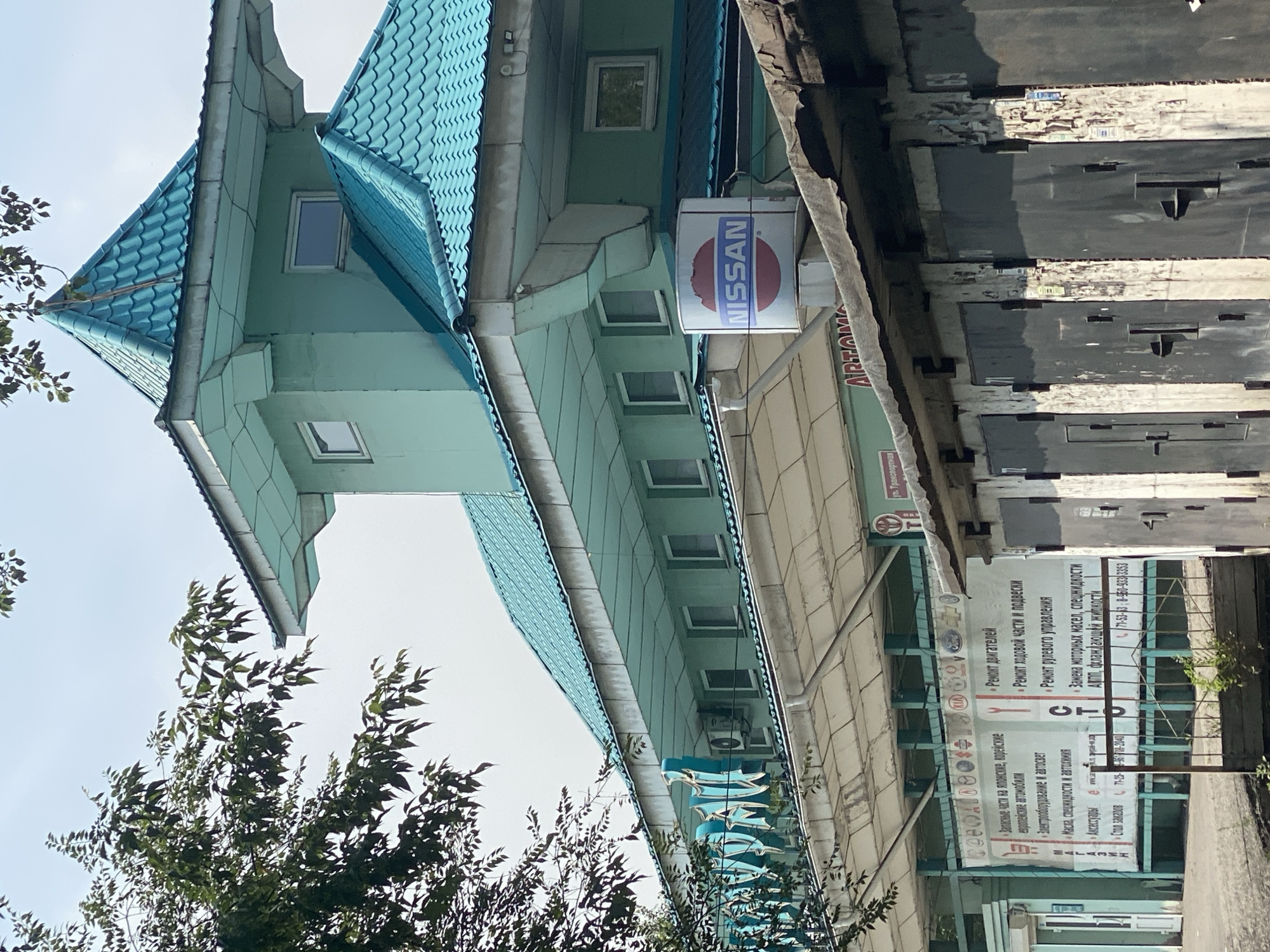 A pale green building with a turquoise tiled roof, resembling traditional East Asian architecture, stands under an overcast sky. In the foreground, a line of dark, weathered garage doors is visible. A circular sign with the "NISSAN" logo is mounted on the side of the building. To the left of the Nissan sign, a blue sign with white lettering is partially visible. Below the building, a large banner displays text and several small brand logos, including Kia and Ford, indicating it is likely an automotive service center. The environment appears to be an urban setting, with trees to the left of the building and a fence in front of the garage doors. The overall impression is one of a commercial establishment, possibly an auto repair shop or dealership, with a distinctive architectural style.