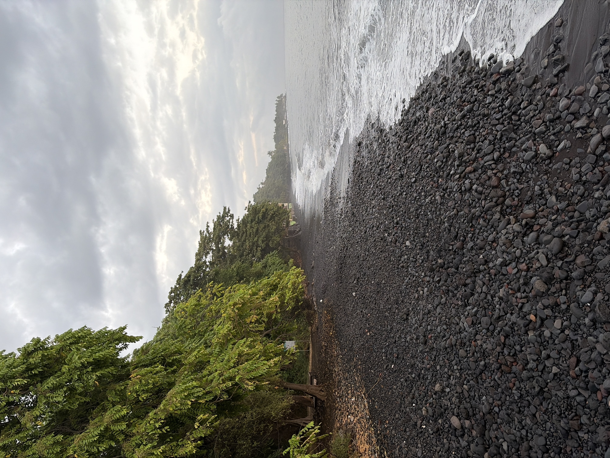 The image captures a volcanic pebble beach in Kubu, Indonesia, under an overcast sky. Lush green trees with dense foliage line the left side of the frame, casting shadows on the dark, rocky shore. The pebbles vary in size and color, predominantly black and gray, with some reddish-brown hues. The ocean water is a muted gray, with gentle waves breaking on the shore, leaving white foam. In the distance, along the coastline, a dense line of trees and some buildings are visible, suggesting a populated area. The overall atmosphere is serene and natural, with a hint of the volcanic origin of the landscape. There are no people or animals visible in the image. The lighting is soft and diffused due to the cloudy sky.