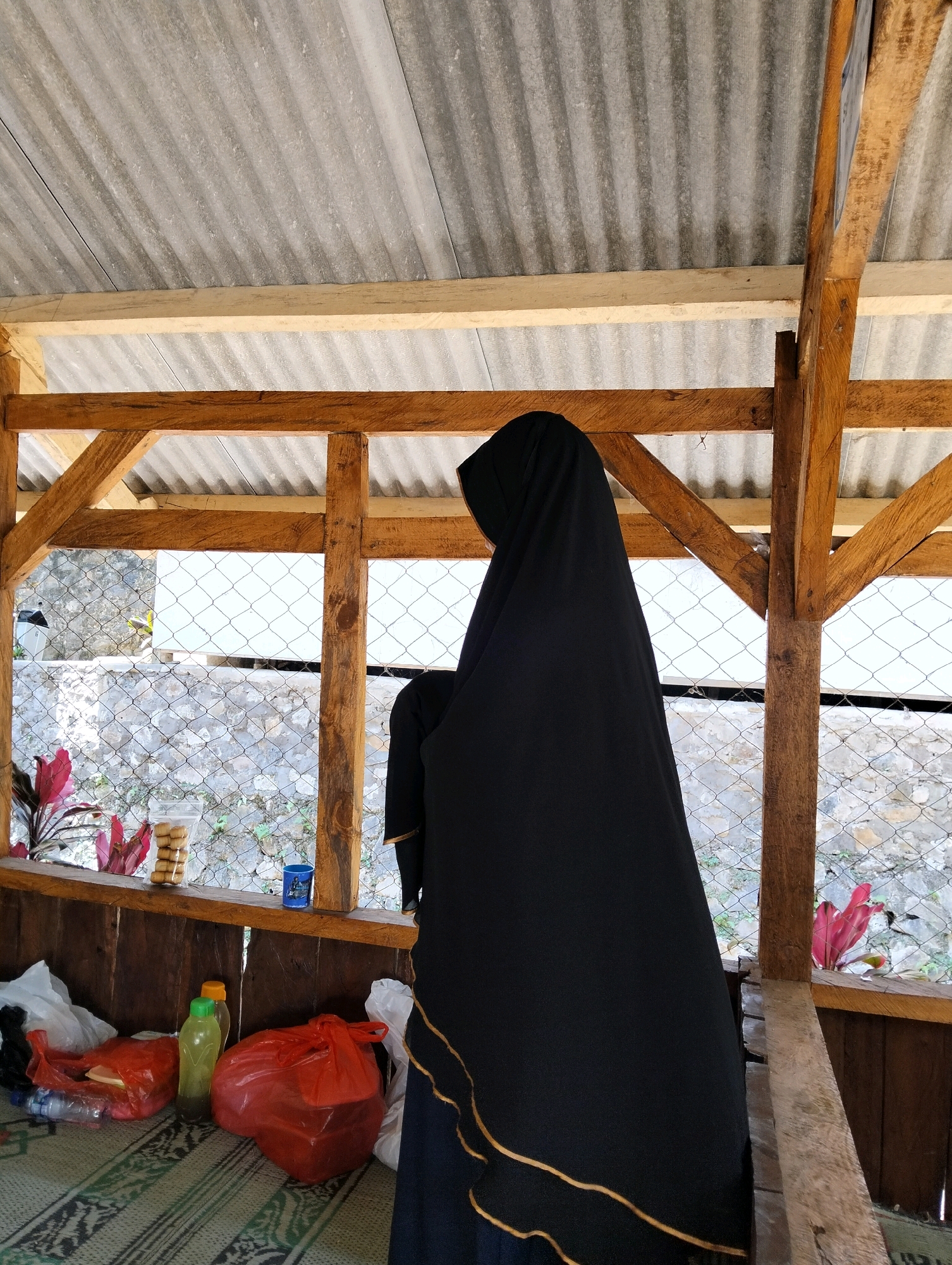 A person wearing a black hijab is standing on a woven mat inside a wooden structure with a corrugated metal roof. In the foreground, there are several plastic bags and bottles on the mat. A narrow shelf runs along the back of the structure, holding a bag of cookies, a blue cup, and a red plastic bag. Through a wire fence behind the shelf, a stone wall and some plants are visible. The wooden beams of the structure are rough and unfinished. The lighting suggests an overcast day. The scene is likely a temporary outdoor shelter, possibly a rest area or a food stall, given the presence of food items. It is located in a somewhat rural or semi-developed area of Subang, Indonesia, indicated by the building materials and the background landscape.