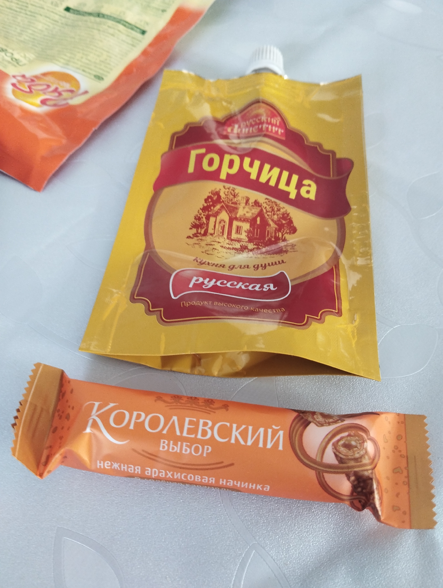 The image depicts a tabletop scene. There is a packet of mustard with Russian labeling and a picture of a house on it, a package of orange dessert bar with Russian labeling that indicates that the item is peanut filled, and a third package that is slightly out of focus in the top left of the image. It is not possible to place this scene in Krasnogorskoye, Russia, based on the image.