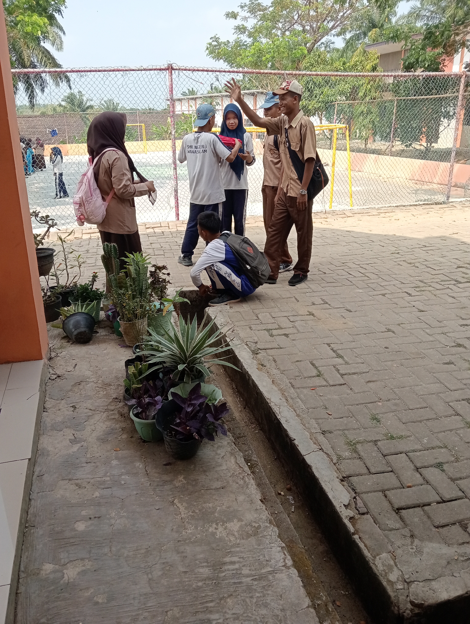 The image depicts an outdoor scene in what appears to be a schoolyard or a similar educational institution in Wanasalam, Indonesia. In the foreground, several potted plants are arranged on a concrete and brick paved area. The plants include cacti, succulents, and dark-leafed varieties. A small drainage ditch runs along the edge of the paved area.
In the mid-ground, a group of four young people, likely students, are gathered near a chain-link fence. Three individuals are wearing school uniforms. One person in a tan uniform is wearing a cap and smiling, with their right arm raised as if waving. Another person in a similar tan uniform stands beside them, also wearing a cap. A third person, wearing a grey t-shirt with text, is looking towards the person in the tan uniform, holding a red object. The fourth person, wearing a hijab, is standing to the left, facing away from the camera, and is carrying a light-colored backpack.
In the background, behind the fence, there is a sports field, possibly a basketball court or similar, with yellow posts visible. Beyond the field, trees and buildings can be seen. The sky is clear and bright, suggesting daytime. A few other figures, possibly students or staff, are visible in the far background on the sports field. The overall impression is of a casual gathering or interaction among students in a school environment.