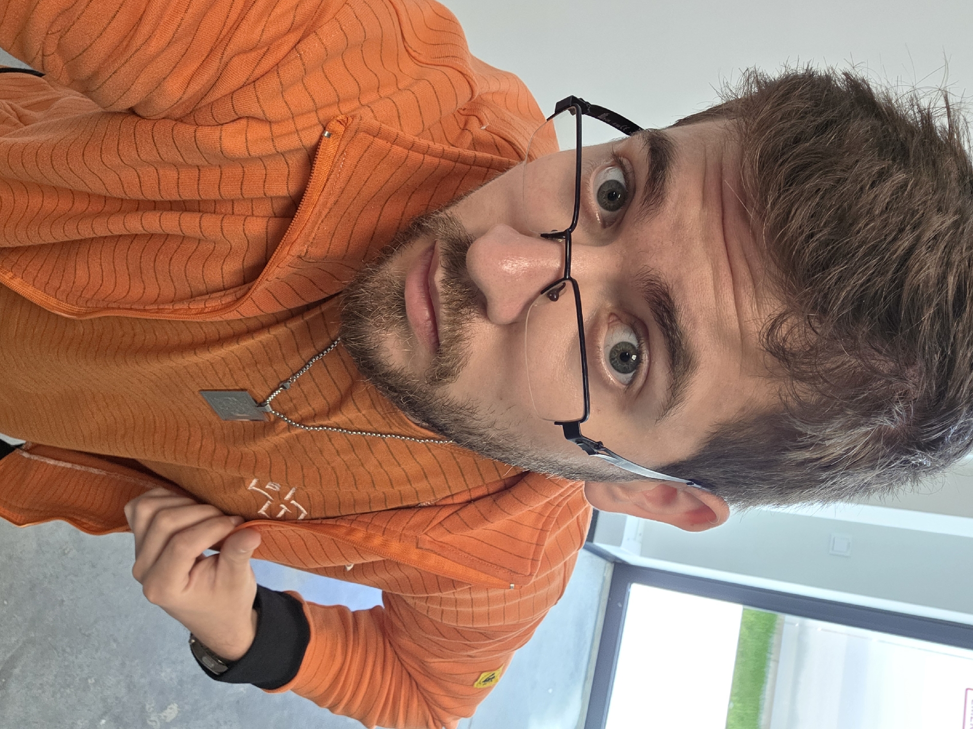 A man wearing an orange jacket and glasses looks at the camera. He has a goatee and a necklace. He is indoors, possibly near a window showing green grass. There is no indication that this is Plovdiv, Bulgaria.