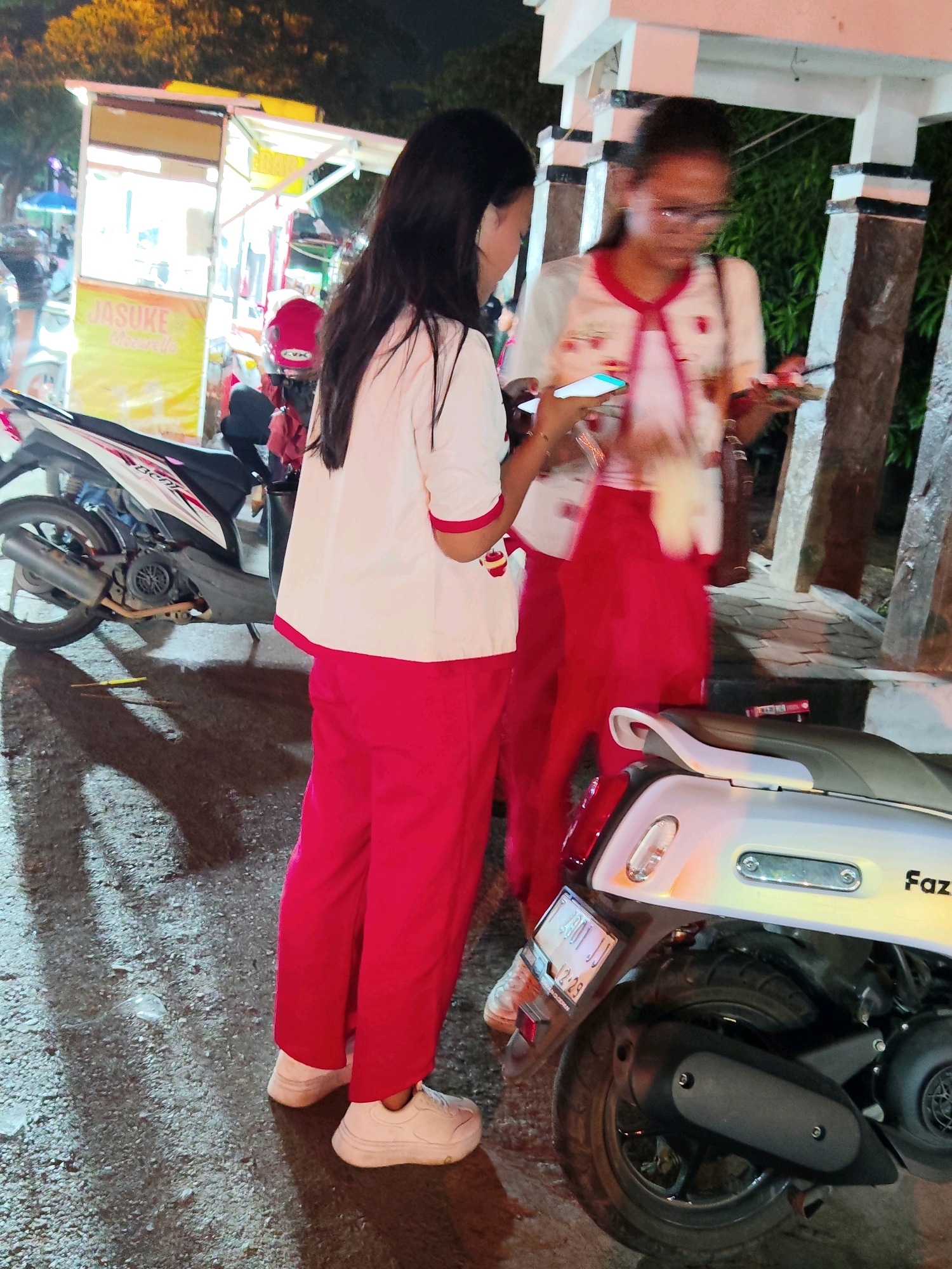 In Purwakarta, Indonesia, two young women in matching red pants and white shirts with red trim stand near a food cart at night. The woman on the left holds a smartphone, and the woman on the right holds a plate with food. A red scooter is parked to the right of the women, and a white scooter is parked to the left. The food cart, labeled "JASUKE Mozzarella," is illuminated and appears to be open for business. The ground is wet and reflective, suggesting it may have rained recently. The overall scene depicts a casual, evening street food encounter in a bustling urban environment.