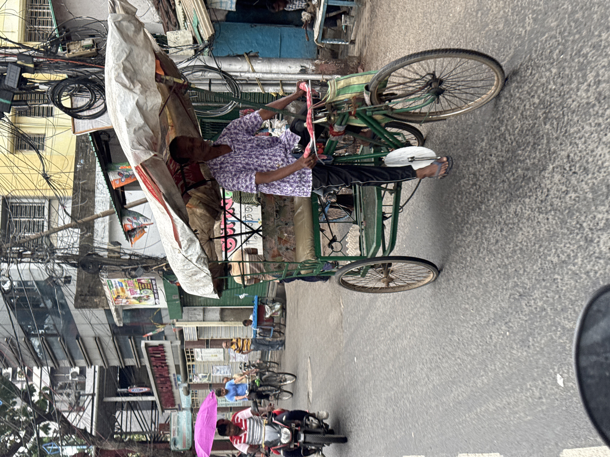A rickshaw driver pedals through a street in Kolkata, India. He wears a lilac patterned shirt and black pants. The rickshaw has a white canopy, and the tires are thin and spoked. The street is lined with buildings, some adorned with intricate wiring and cables. Motorbikes and pedestrians share the road with the rickshaw. The lighting is overcast, and the road surface is a worn gray.