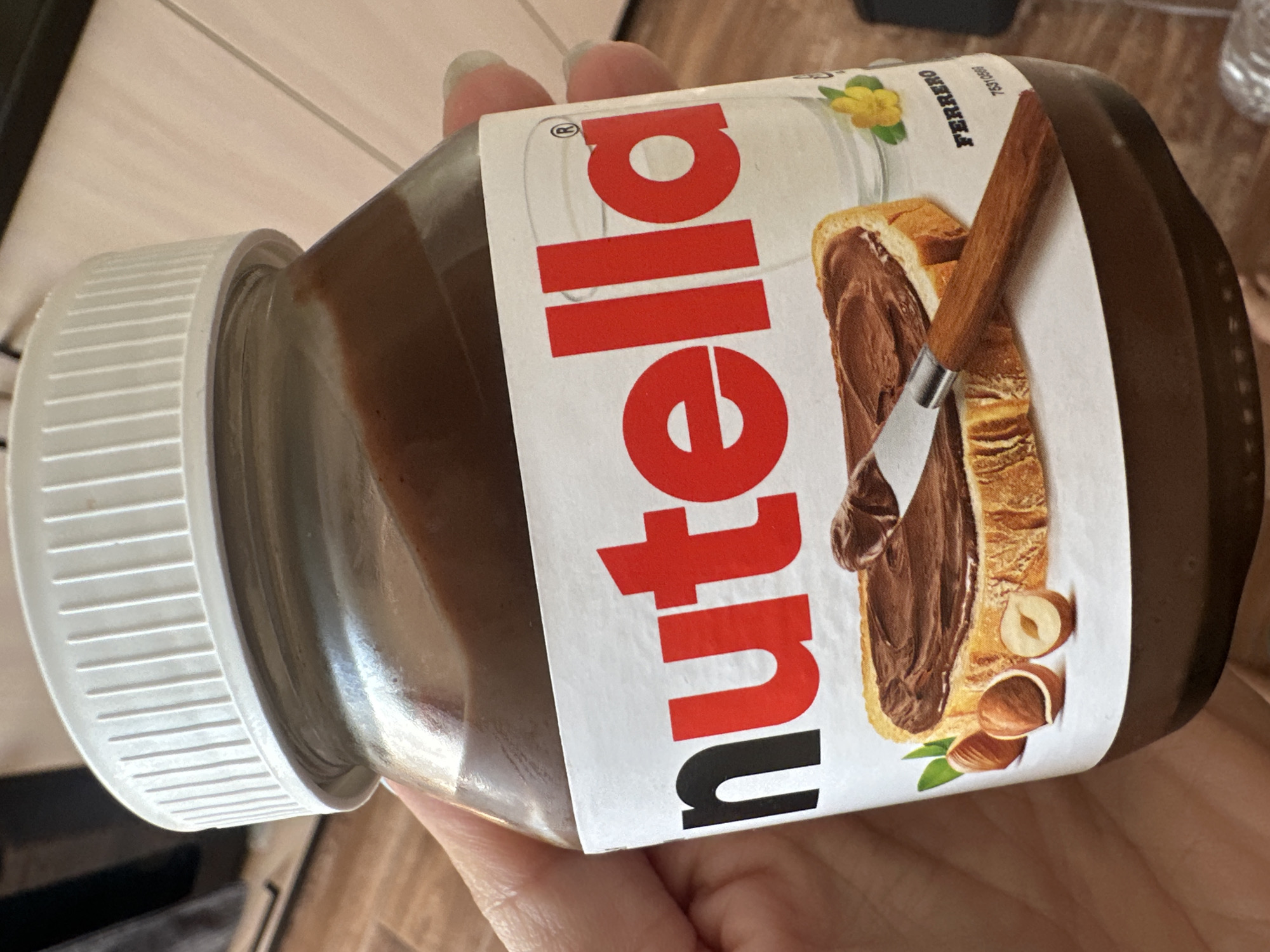 An indoor scene shows a person's hand holding a glass jar of Nutella. The jar has a white lid and a label that prominently displays the "Nutella" logo in red and black lettering. The label also features an illustration of a slice of bread with Nutella spread on it, along with hazelnuts and a yellow flower. The background is blurred, but suggests a wooden surface and a dark object. The scene does not provide any information about the location being in Novoaltaysk, Russia.