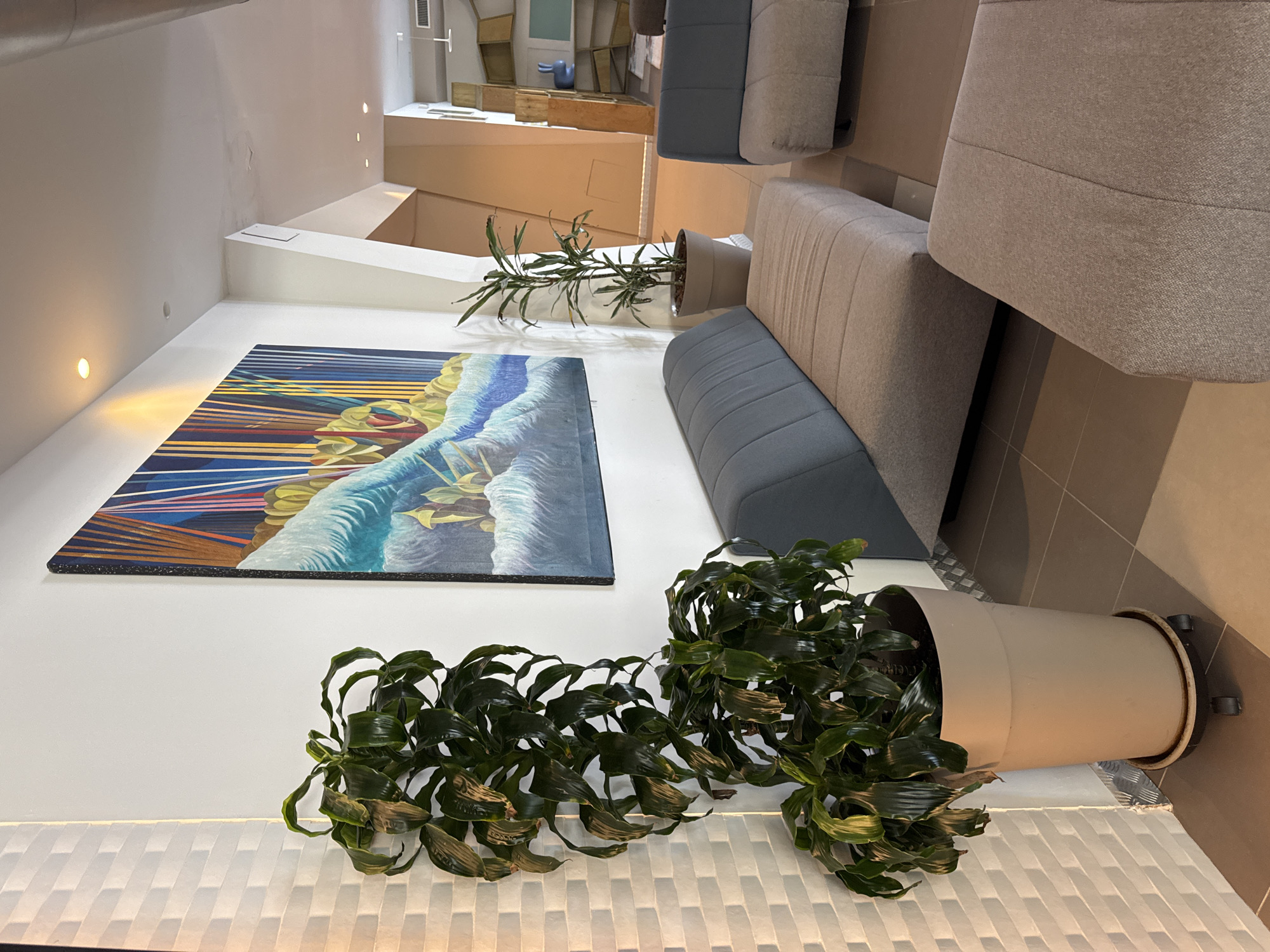 This image depicts a modern, minimalist interior space, likely a waiting area, lounge, or hotel reception, situated in Isola delle Femmine, Italy. The room features clean lines, neutral colors, and contemporary furnishings.
On the main wall, a large, vibrant painting is prominently displayed. It is a semi-abstract piece depicting dynamic blue and white waves or water, set against a background of striking yellow, red, and dark blue linear patterns, possibly representing a dramatic sky or light effects. Below the painting, a low, textured white wall or bench serves as a base for two large potted plants. These plants have dark green, broad leaves, some of which show signs of browning or wilting. They are housed in tall, light-colored, cylindrical planters.
To the right, a series of modern, modular sofas or benches are arranged. These seating units are upholstered in shades of grey and light beige fabric, featuring simple, clean designs. The floor is tiled in a neutral tone that complements the furniture.
The room is illuminated by warm recessed lighting embedded in the walls, creating a soft glow. In the background, a multi-tiered reception desk or counter in a light, possibly wooden or laminate, material is visible. Further back, an open shelving unit with various compartments can be seen, and a small, abstract blue sculpture or decorative object rests on one of the shelves.
No people are present in the image, and there is no visible activity taking place. The scene conveys a sense of quiet and modern sophistication. No discernible text is visible within the image.