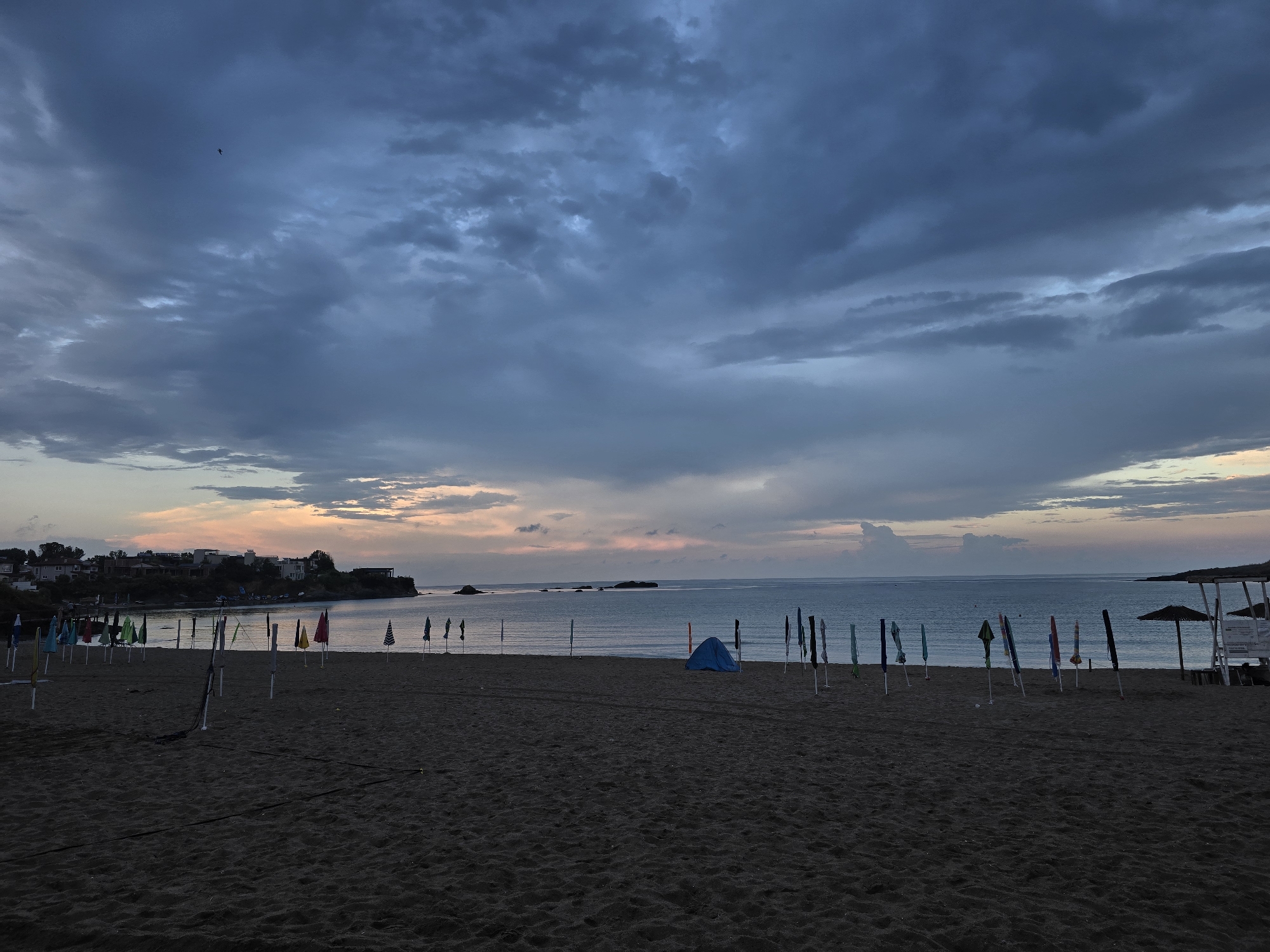 The image depicts a beach scene at dusk. The sky is filled with dramatic, dark clouds, with streaks of soft orange and pink light visible near the horizon. A single bird is silhouetted against the cloudy sky. The beach itself is dark sand, with numerous closed beach umbrellas and their poles scattered across the foreground and middle ground. In the middle ground, a small blue tent is pitched on the sand, and a few beach chairs are visible further to the right. The sea is calm and reflects the muted colors of the sky. In the background, a coastline with buildings is visible on the left, and a rocky promontory extends into the sea on the right. The overall impression is one of a quiet, approaching evening on a deserted beach.