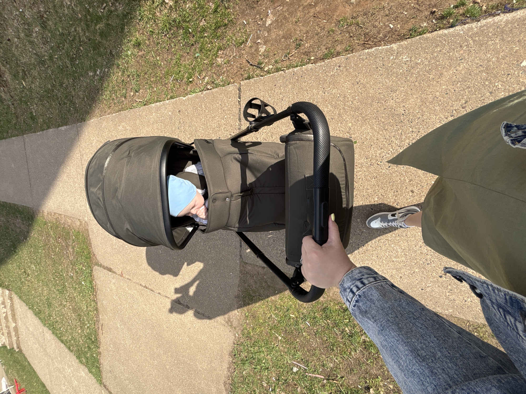 A person is pushing a baby in a stroller on a concrete sidewalk adjacent to a patch of grass. The baby is wearing a blue hat and lying in the stroller. The person pushing the stroller is wearing a denim jacket and olive green pants, with grey sneakers visible. The stroller is dark green and has a black handle. The scene appears to be outdoors on a sunny day. The location is not discernible from the image.