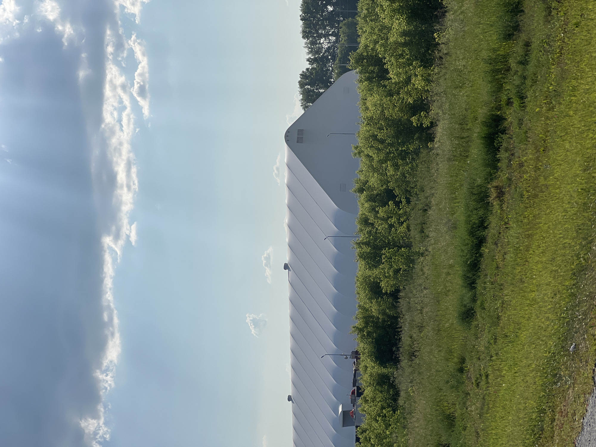 A large, white, arched structure, likely an industrial tent or arena, dominates the midground of the image. It is situated in Verkh-Tula, Russia. The structure is covered with a white, taut fabric that displays a repeating pattern of vertical pleats or segments. Two dark, circular vents are visible on the roof of the structure towards the left side. Towards the left edge of the structure, there are several smaller objects, including a white trailer with an orange tarp on top and what appear to be vehicles. Several light poles are positioned in front of the structure.
The structure is surrounded by dense, green foliage and trees. In the foreground, there is a field of tall, green grass and other vegetation, with some areas appearing more sunlit than others. In the background, behind the white structure, a line of dark green trees forms a backdrop. The sky above is a light blue, with scattered white and gray clouds. Sunlight streams through the clouds, creating visible rays. The overall scene appears to be an outdoor environment on a sunny day. There are no visible people in the image.