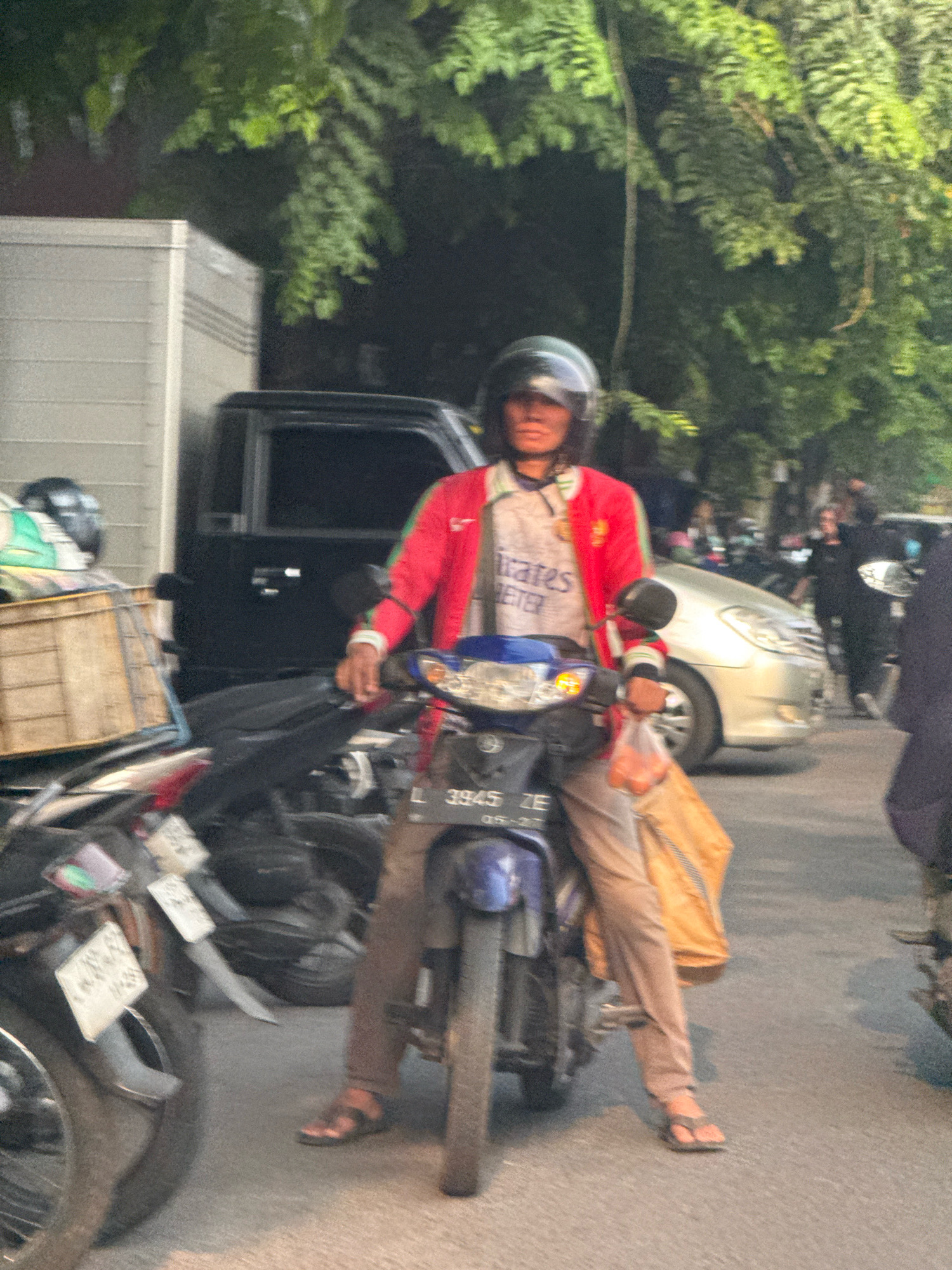 The image depicts a street scene in Surabaya, Indonesia, during daylight. In the foreground, a person is seated on a motorcycle, wearing a red jacket with green accents and a light-colored shirt underneath. The person has on a helmet and appears to be holding a plastic bag with food items. The motorcycle has a blue front panel and a license plate with the characters "L 3945 ZE". Behind the motorcycle, other vehicles, including a silver car and several other motorcycles, are visible. A large white truck is parked to the left of the scene. Trees with green foliage line the street, indicating an outdoor, urban environment. The overall impression is a snapshot of everyday traffic and urban life in Surabaya.