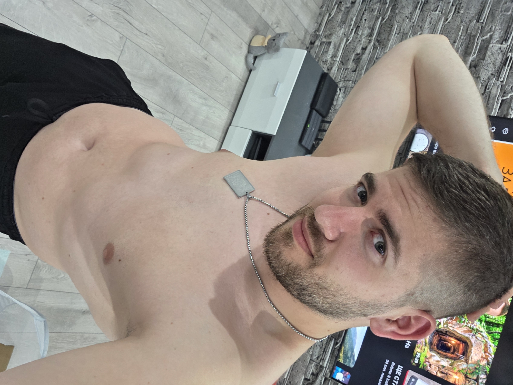 A torso of a shirtless man wearing black shorts and a silver dog tag necklace is shown. He has a beard and short brown hair. He is indoors, standing in front of a television and other furniture. The television shows a video of a cave or tunnel. The floor is light grey wood laminate, and the wall behind the TV is tiled with a dark grey textured tile. A stuffed mouse toy sits on top of a white printer or scanner. The location is not discernible from the image.