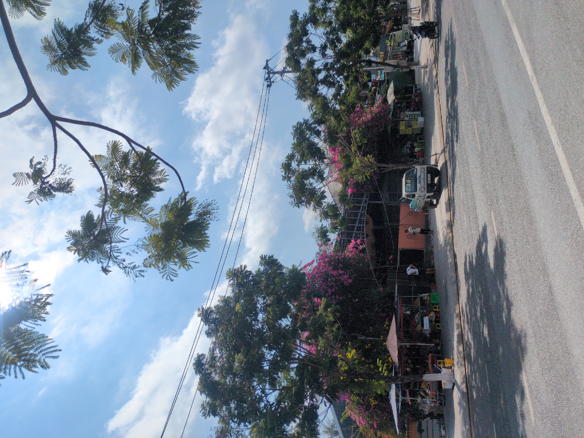 The image shows a street scene in Phú Quốc, Vietnam. A paved road runs along the right side of the frame. A white utility truck is parked on the roadside. A storefront or market stall is visible near the truck. Several people are observed near the stall. Trees line the street, with flowering bougainvillea visible. Overhead power lines traverse the sky, which is partly cloudy.