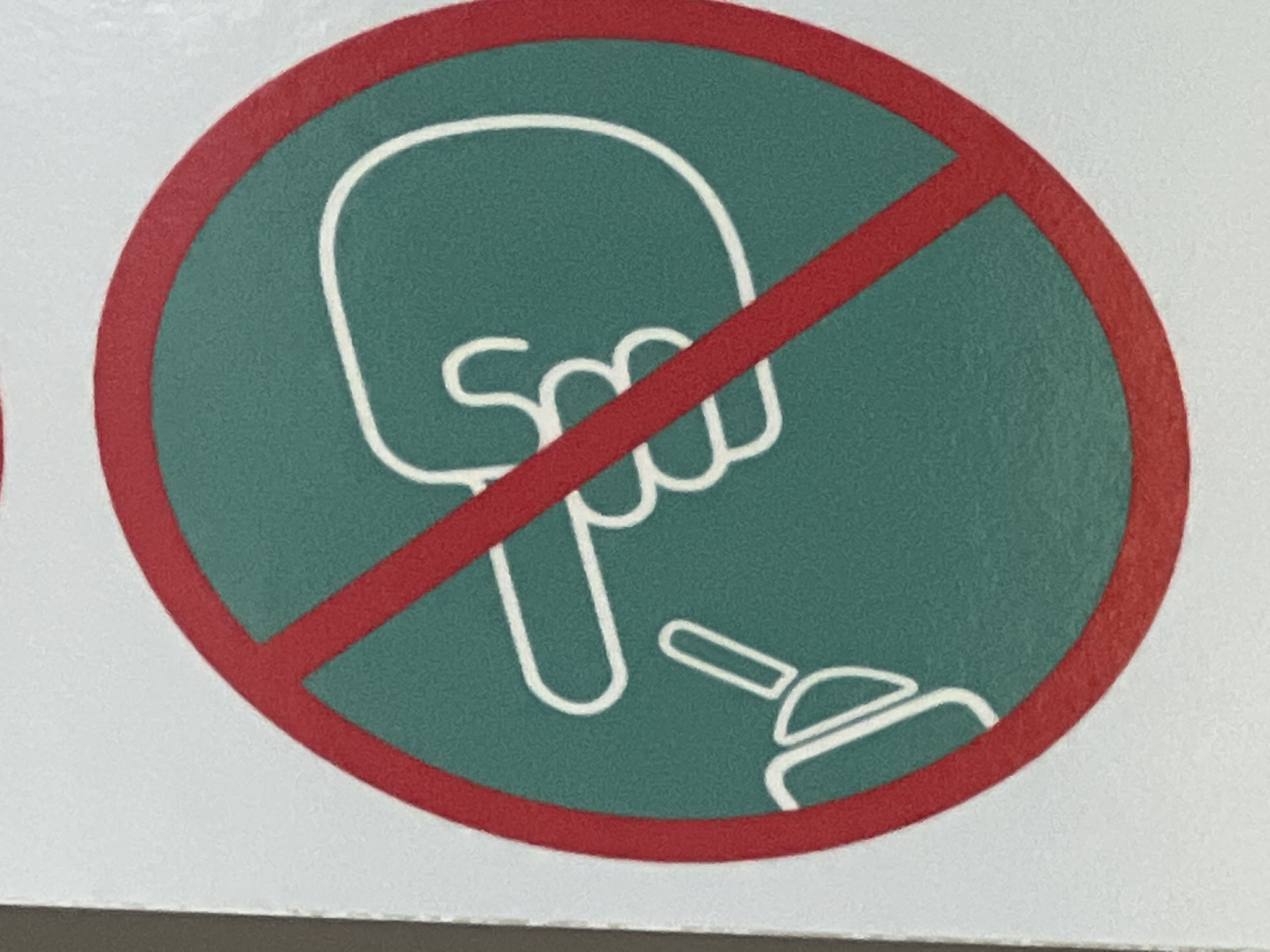 This is a close-up, top-down, slightly angled, eye-level shot of a circular prohibition sign. The sign has a thick red border and a teal green background. A thick red diagonal line crosses the circle from the top left to the bottom right, indicating prohibition. Inside the circle, a white outline depicts a human hand with its index finger pointing down towards a stylized object that resembles a light bulb with a cap. The hand is shown from a perspective that suggests it is about to touch or interact with the object. The image lacks any context of its specific location within Vilnius, Lithuania, or any surrounding environment.