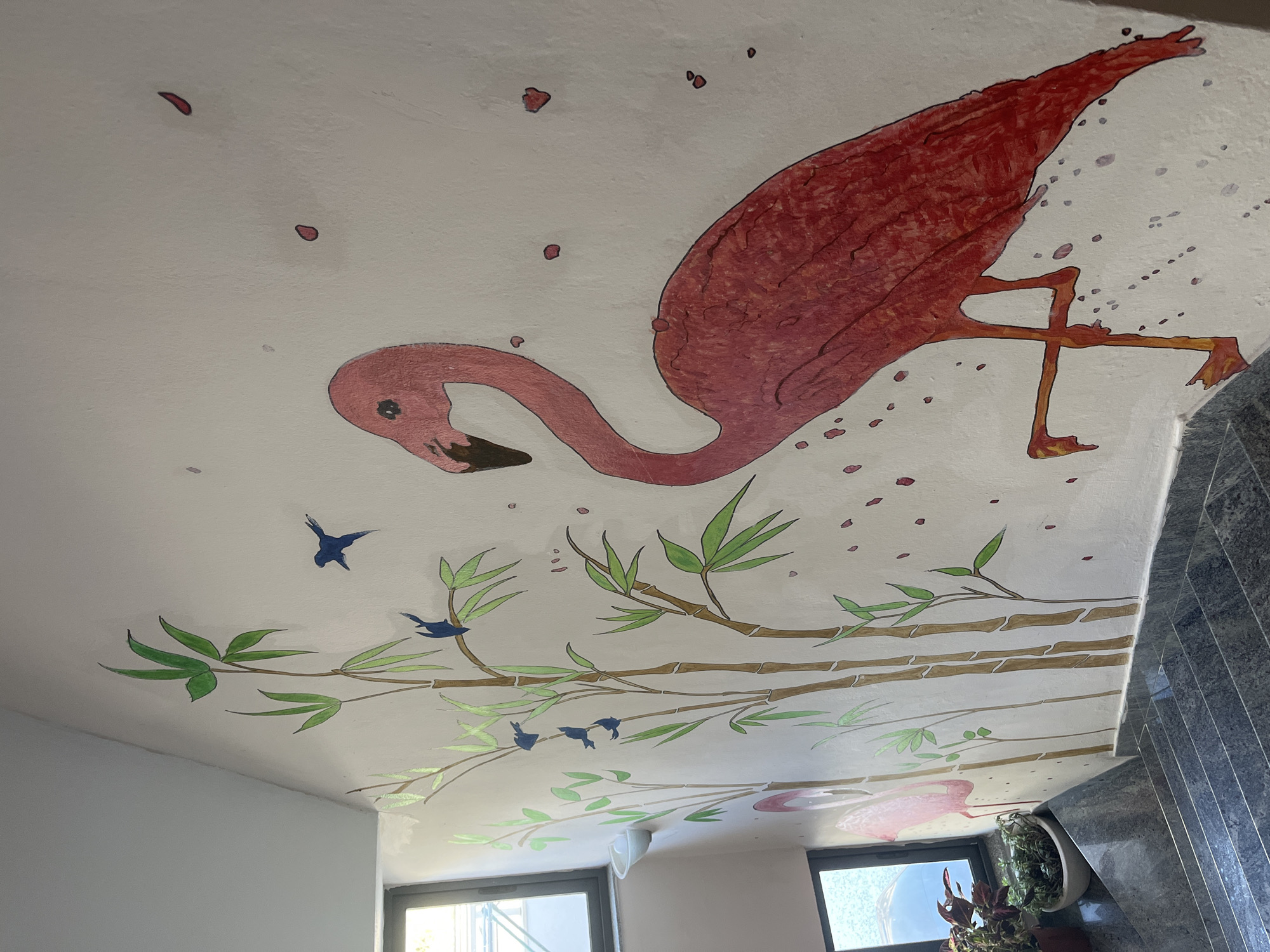 A mural adorns the wall of a stairwell in Bashkia Durrës, Albania. The artwork depicts a large, vibrant pink flamingo standing on one leg, its neck gracefully curved. To the left of the flamingo, a cluster of bamboo stalks with green leaves rises up the wall. Several small, dark blue birds are painted on the bamboo branches. The wall itself is white, with scattered pink splotches that resemble falling petals or paint splatters. Below the mural, a dark gray granite staircase descends. Two windows are visible on the left side of the stairwell, one higher than the other, with natural light filtering in. Potted plants are placed on the windowsill of the lower window. The overall scene suggests a decorative element within a building, likely a home or public space.