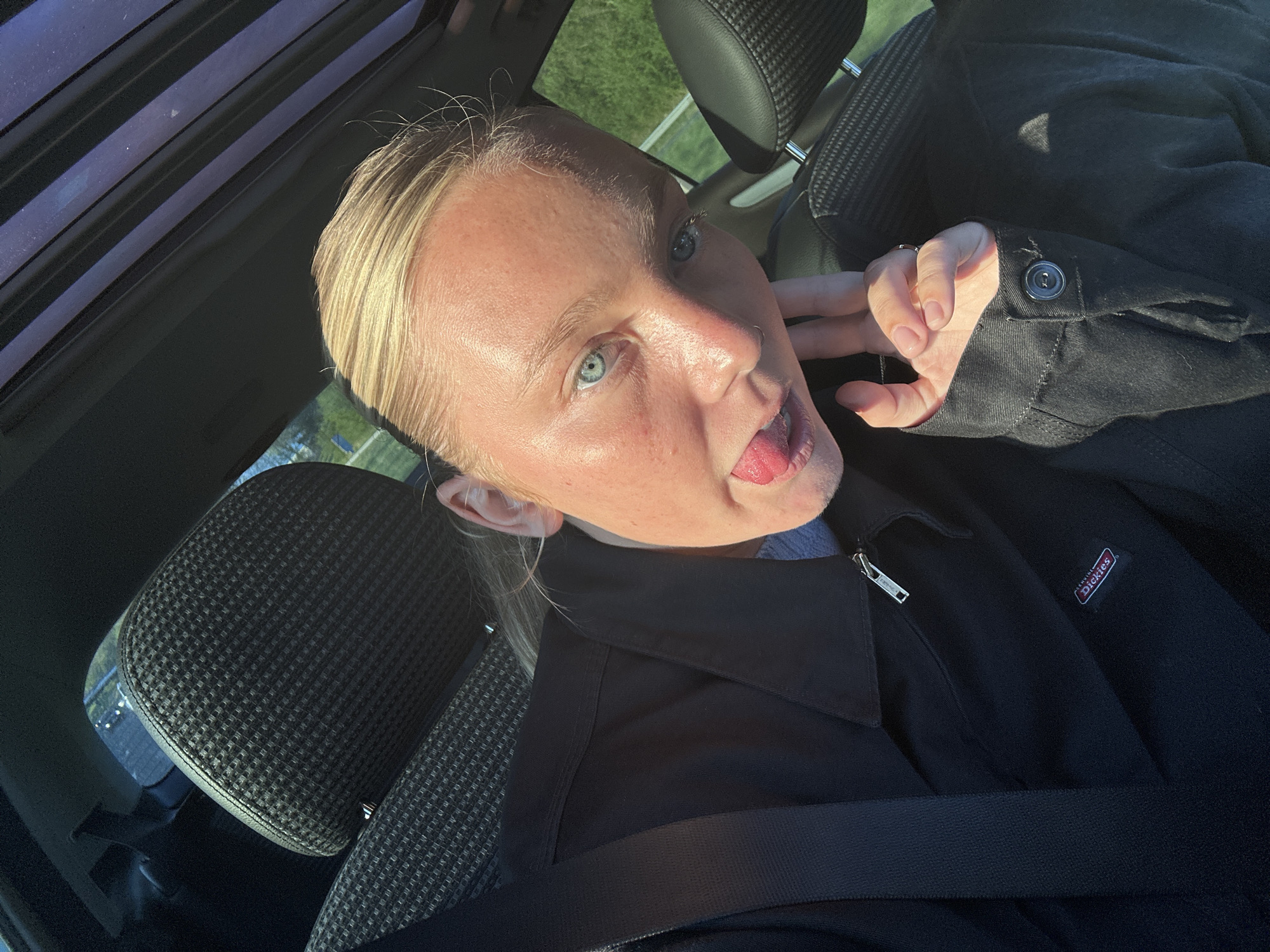 A light-skinned individual with blonde hair pulled back is in the driver's seat of a car. They are sticking their tongue out and making a face, with their hand near their cheek. They are wearing a black jacket with a "Dickies" label and a visible seatbelt. The car has a sunroof. The interior has black headrests with a textured pattern. The location, Jossigny, France, cannot be determined from the image.