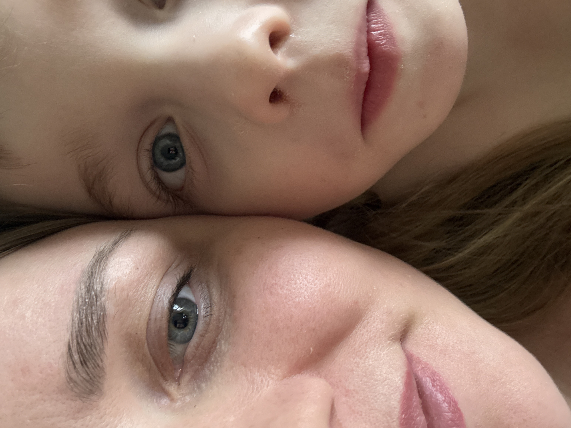 The image shows the faces of two individuals, presumably a child and an adult, likely a mother. Both are fair-skinned with blue eyes. The child's face is positioned above the adult's face, with the two faces aligned closely. The perspective is close-up, focusing on the facial features. The child has fine blonde hair and a small nose. The adult has defined eyebrows and fuller lips. The image's background is blurred, suggesting a shallow depth of field. There is no indication of the location being Chaikovsky, Russia.
