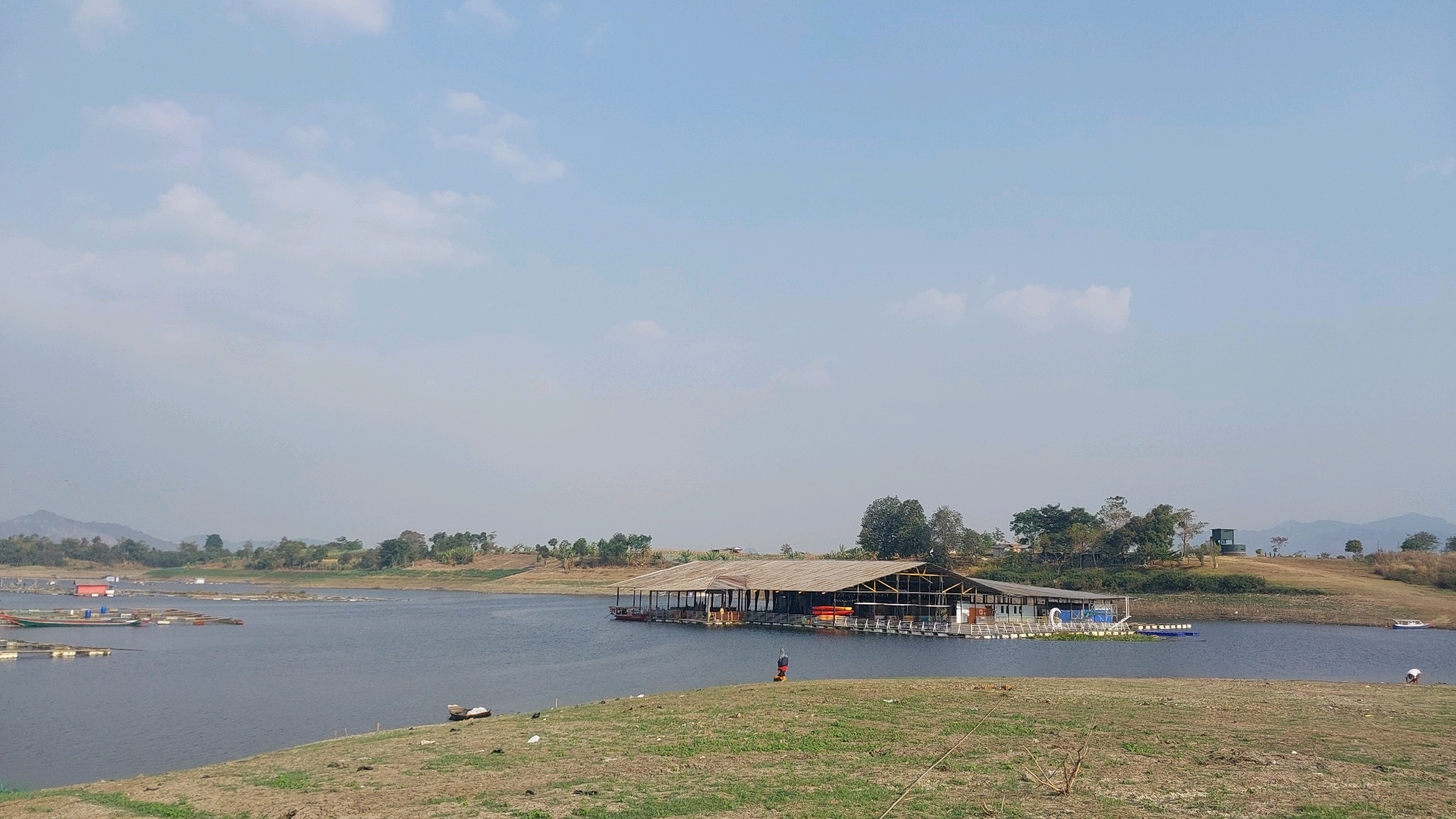 The image depicts a wide, calm body of water, likely a reservoir or lake, under a hazy, overcast sky with sparse clouds. In the middle of the water, a large, long, multi-sectioned floating structure with a dark, thatched roof and open sides is moored. This structure appears to be a restaurant or gathering place, with several smaller boats and what look like aquaculture pens visible in the distance to the left. The shoreline in the foreground is dry, grassy, and uneven, with some scattered debris. The opposite shore is lined with trees and rolling hills, with a few scattered buildings and a distinctive small, green tower-like structure visible on a higher elevation. A single person in an orange shirt is visible standing on the floating structure, and another person in white is visible near the water's edge on the right side of the frame. The scene conveys a sense of rural tranquility. The location is identified as Bojonghaleuang, Indonesia.