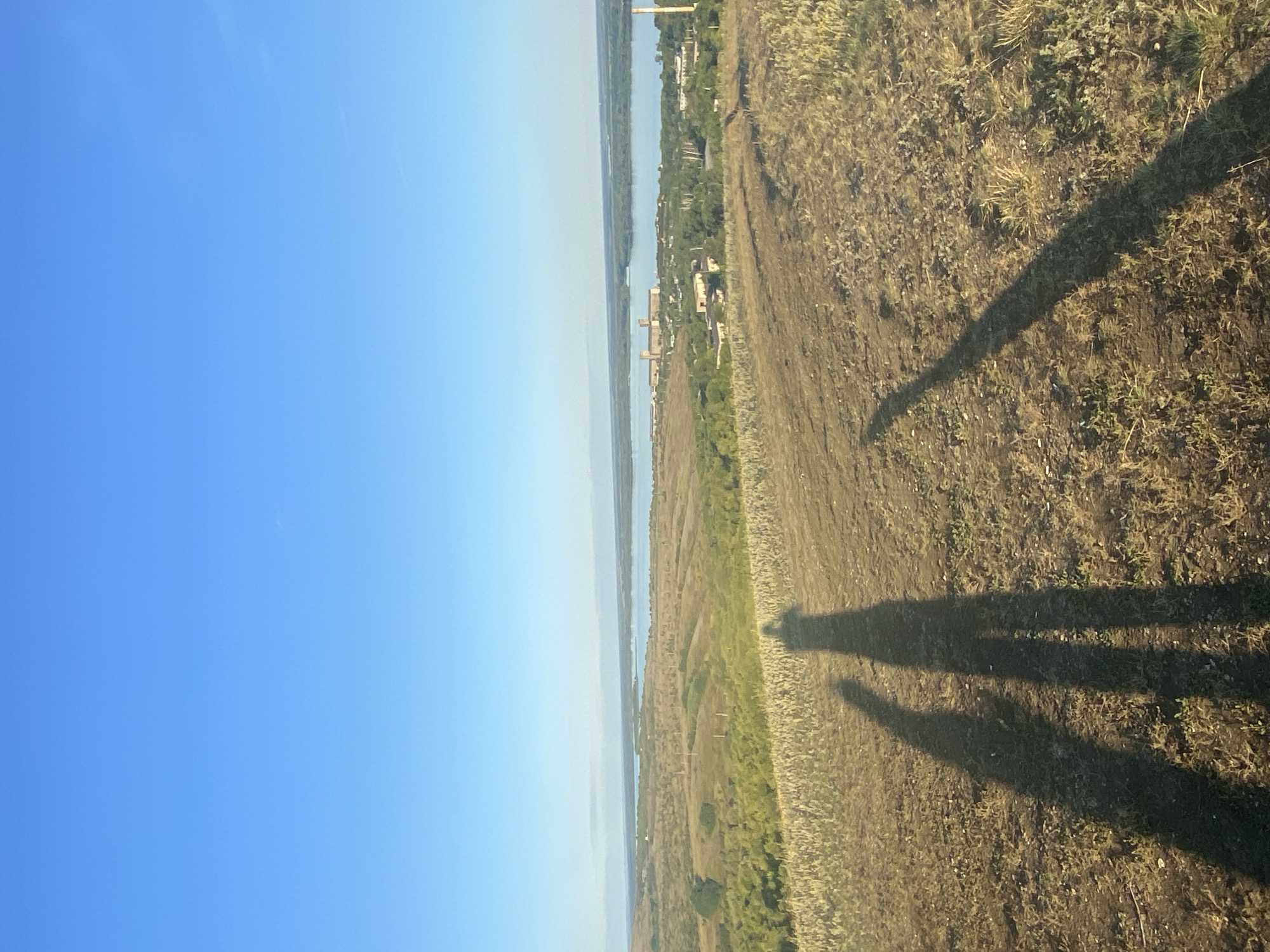 A wide-angle, full-frame photograph captures a pastoral landscape under a clear, bright blue sky. In the foreground, three distinct human shadows are cast diagonally across dry, brown earth dotted with sparse, dry grass. The shadows suggest the presence of at least three individuals standing on a slight incline. Beyond the foreground, the terrain slopes down, revealing a body of water, likely a river or reservoir, that stretches horizontally across the mid-ground. On the opposite bank of the water, a collection of buildings is visible, including industrial structures with smokestacks and smaller residential or administrative buildings. Greenery, consisting of trees and fields, surrounds the water and buildings, indicating a semi-rural or industrial area within a natural setting. The overall impression is a serene, sunlit day with a view overlooking a settlement and natural water feature. This vista is consistent with views from the hills near Oktyabrsk, Russia, which overlooks the Volga River.