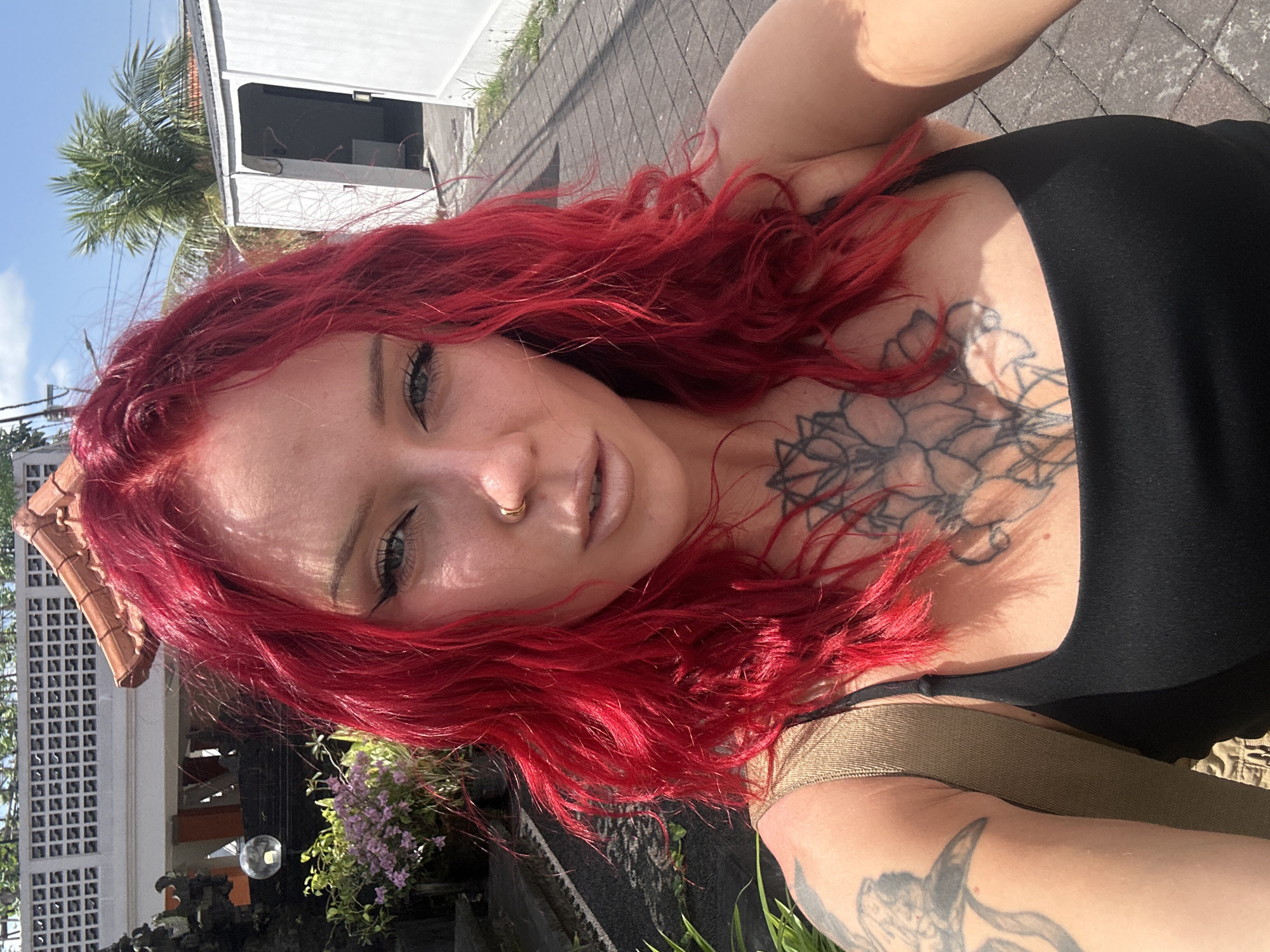 A woman with red hair and tattoos poses for a selfie in North Kuta, Indonesia. She is wearing a black top and a tan strap is visible. The background features tropical plants, a building with a palm tree, and a tiled walkway.