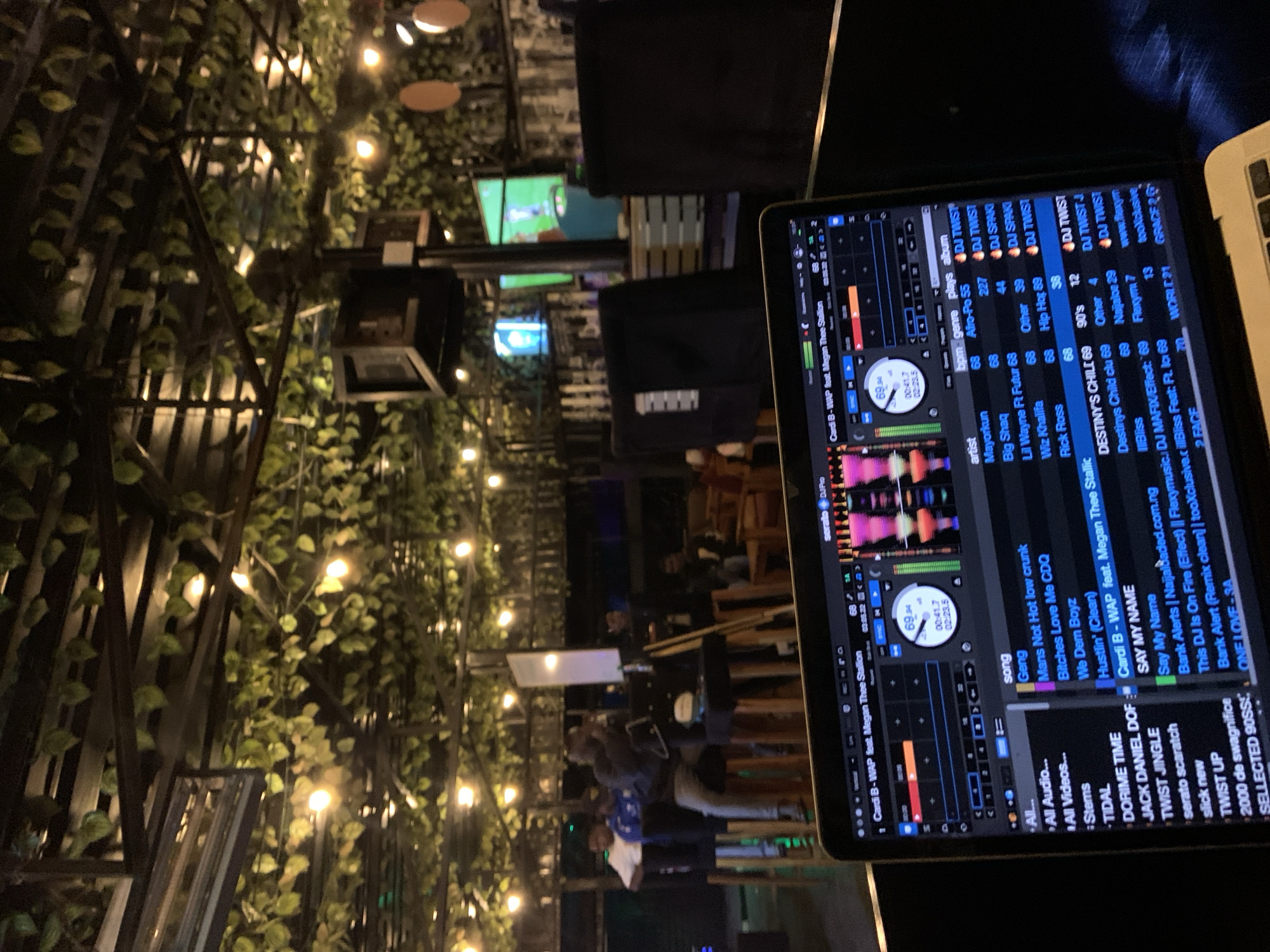This image depicts a dimly lit interior scene, likely a bar or club, with a focus on a laptop displaying DJ software. The laptop screen is prominently in the foreground, showing waveforms, track information, and controls of a DJ program, specifically showing "Cardi B - WAP feat. Megan Thee Stallion" as the current track. In the background, several individuals are visible, some seated and others standing. One person is seated on a high stool, appearing to be speaking into a microphone, while others are gathered around. The environment is decorated with artificial foliage and string lights, creating a cozy and ambient atmosphere. The overall impression is that of a lively entertainment venue. The location is identified as Gura Top, Nigeria.