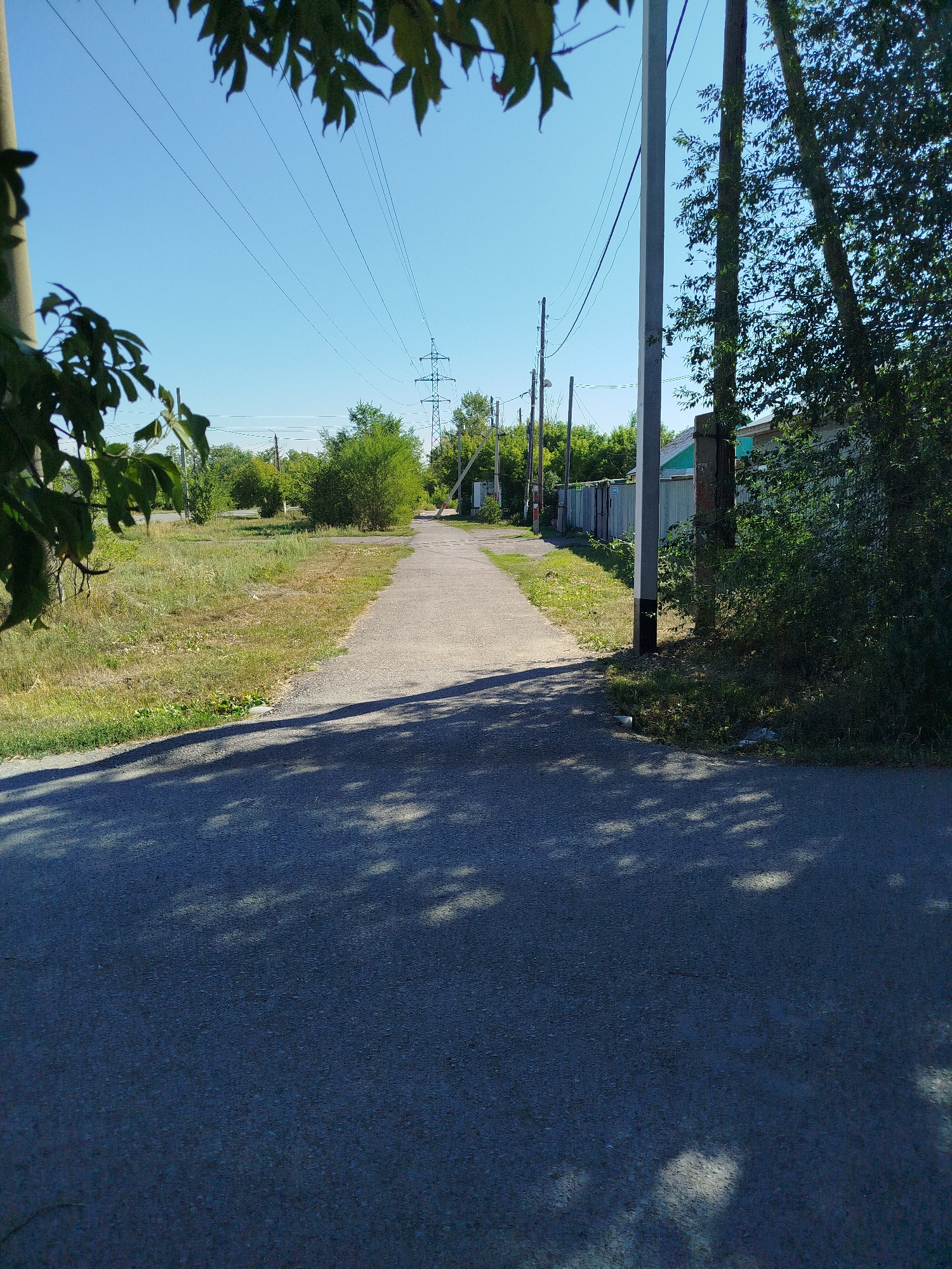 The image depicts a paved road leading into a rural or suburban area. Overhead power lines traverse the clear blue sky, supported by utility poles. Flanking the road are grassy verges, with clusters of green trees and shrubs. On the right side of the road, a metal fence runs along a property line. In the foreground, dappled sunlight casts shadows on the asphalt surface. The scene suggests a quiet, possibly undeveloped area within or on the outskirts of Qarağandı city administration, Kazakhstan.