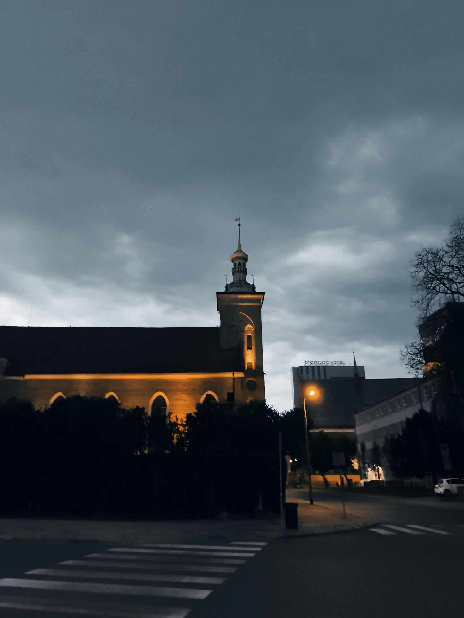 The image depicts an evening street scene in the city of Gdańsk, Poland, likely near the historic Old Town. A prominent church with a lit tower stands on the left, its arched windows glowing with warm light. In the background, a modern building with the "Mercure HOTEL" sign is visible. The sky is overcast and dark, suggesting twilight or approaching night. A street lamp casts an orange glow onto the wet asphalt of the road. A pedestrian crossing is marked on the road in the foreground. Trees and foliage partially obscure the lower sections of the buildings. A white car is parked on the right side of the street. The overall atmosphere is moody and atmospheric, with a contrast between the old and new architecture.