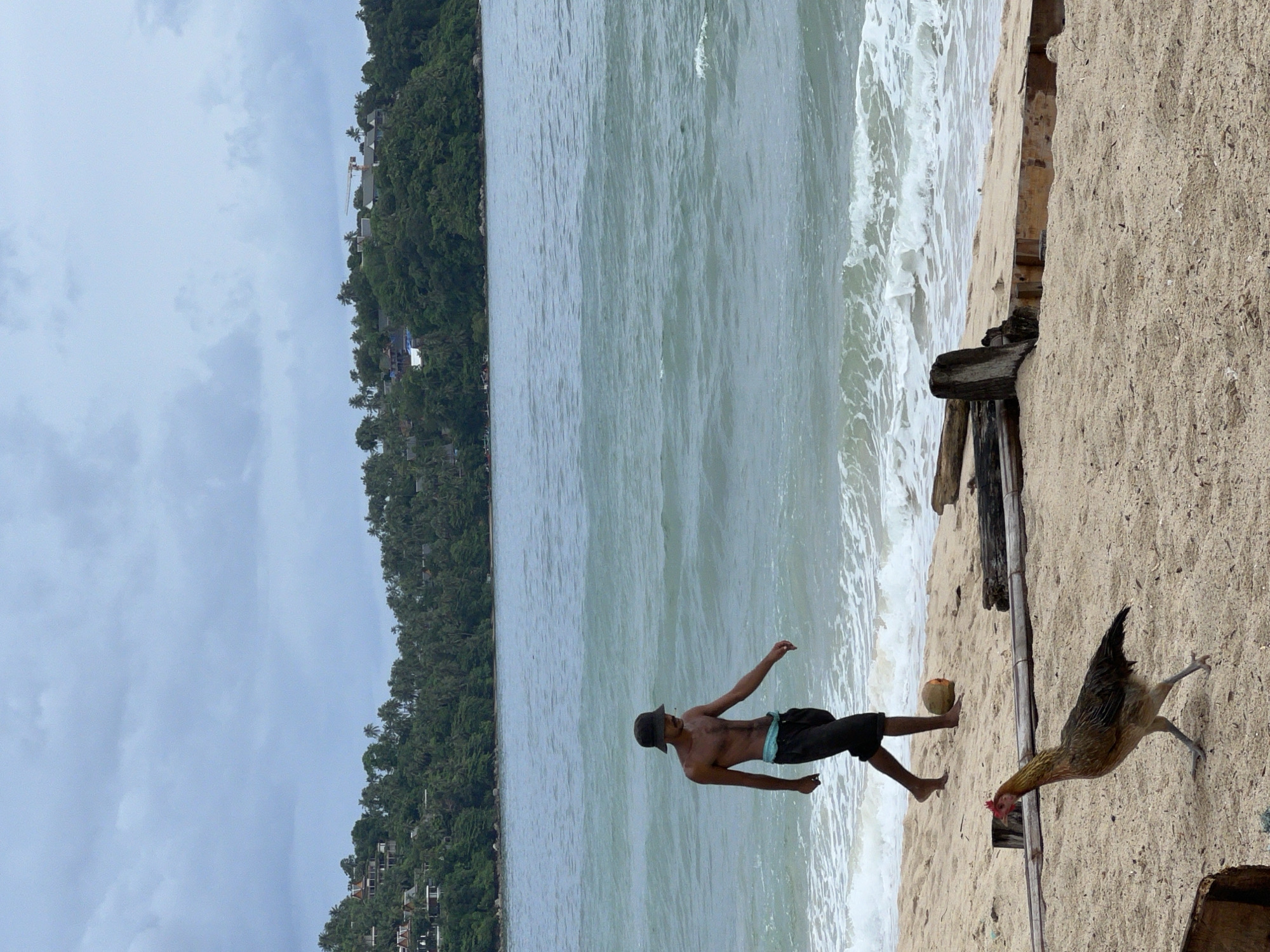 A man wearing a bucket hat and dark shorts walks on a sandy beach towards the ocean. He is shirtless and appears to be kicking a coconut. A chicken walks on the sand in the foreground, facing left. Behind the man, the ocean waves crash on the shore under a cloudy sky. Lush green hills with buildings are visible in the distance across the water. Wooden debris is scattered on the beach.