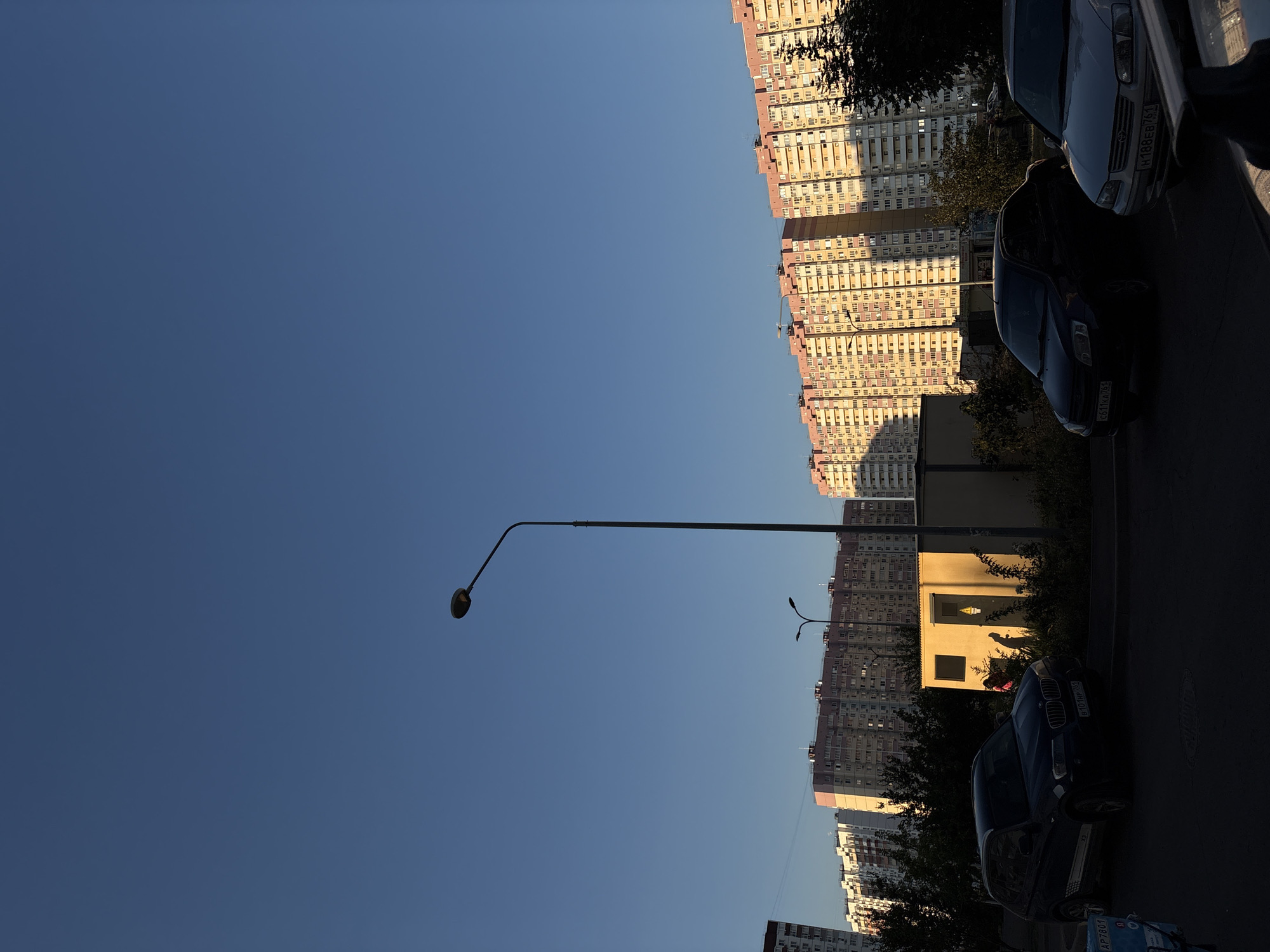 The image depicts a street scene with residential buildings and parked cars under a clear blue sky. In the foreground, several cars are parked, with a dark-colored BMW X3 on the left and a dark sedan and a silver sedan further to the right. Behind the cars, there's a small, yellow, rectangular building with a window and a door, casting a strong shadow. Two figures, silhouetted against the bright light, are near this building, with one appearing to be wearing a pink garment.
In the background, a series of tall, multi-story apartment buildings with light-colored facades and pink roofs dominate the skyline. These buildings appear to be modern residential blocks. A street lamp is visible in the middle ground, and a taller street lamp is positioned to the left of the apartment buildings. The lighting suggests it is late afternoon or early evening, with the sun casting long shadows. The environment appears to be a typical urban residential area.
Based on the license plates visible on the cars, specifically "B101HP761" on the BMW and "H188EB761" on the silver sedan, the vehicle registration codes indicate the Rostov region of Russia. The "761" is the code for Rostov-on-Don. This suggests the scene is located in or around the city of Rostov-on-Don, Russia.