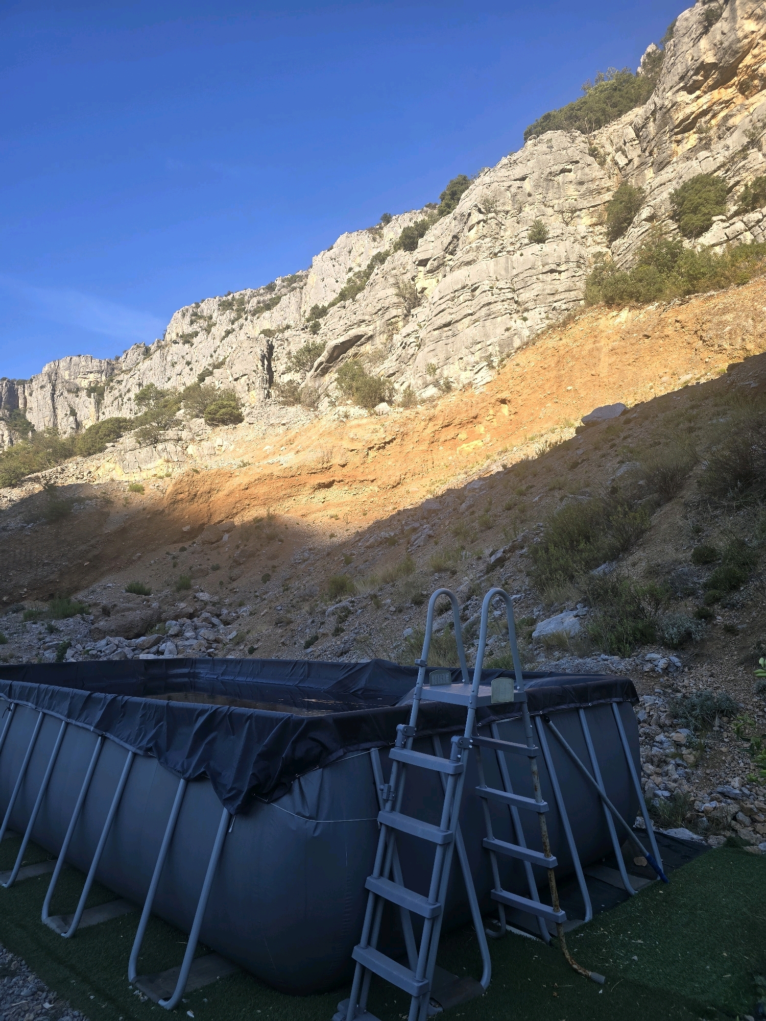 The image depicts an outdoor scene featuring an inflatable swimming pool set against a dramatic natural backdrop. The pool, dark gray with a black cover, is supported by silver-colored metal poles. A gray ladder with multiple steps is attached to the pool. The surrounding ground is covered with artificial green turf and rough terrain with scattered rocks. Behind the pool, a steep, rocky hillside dominates the landscape, characterized by layered rock formations and patches of reddish-orange soil. Sparse vegetation, including bushes and small trees, is visible growing on the slopes. The sky above is a clear, bright blue. The scene appears to be situated in a rugged, mountainous area.