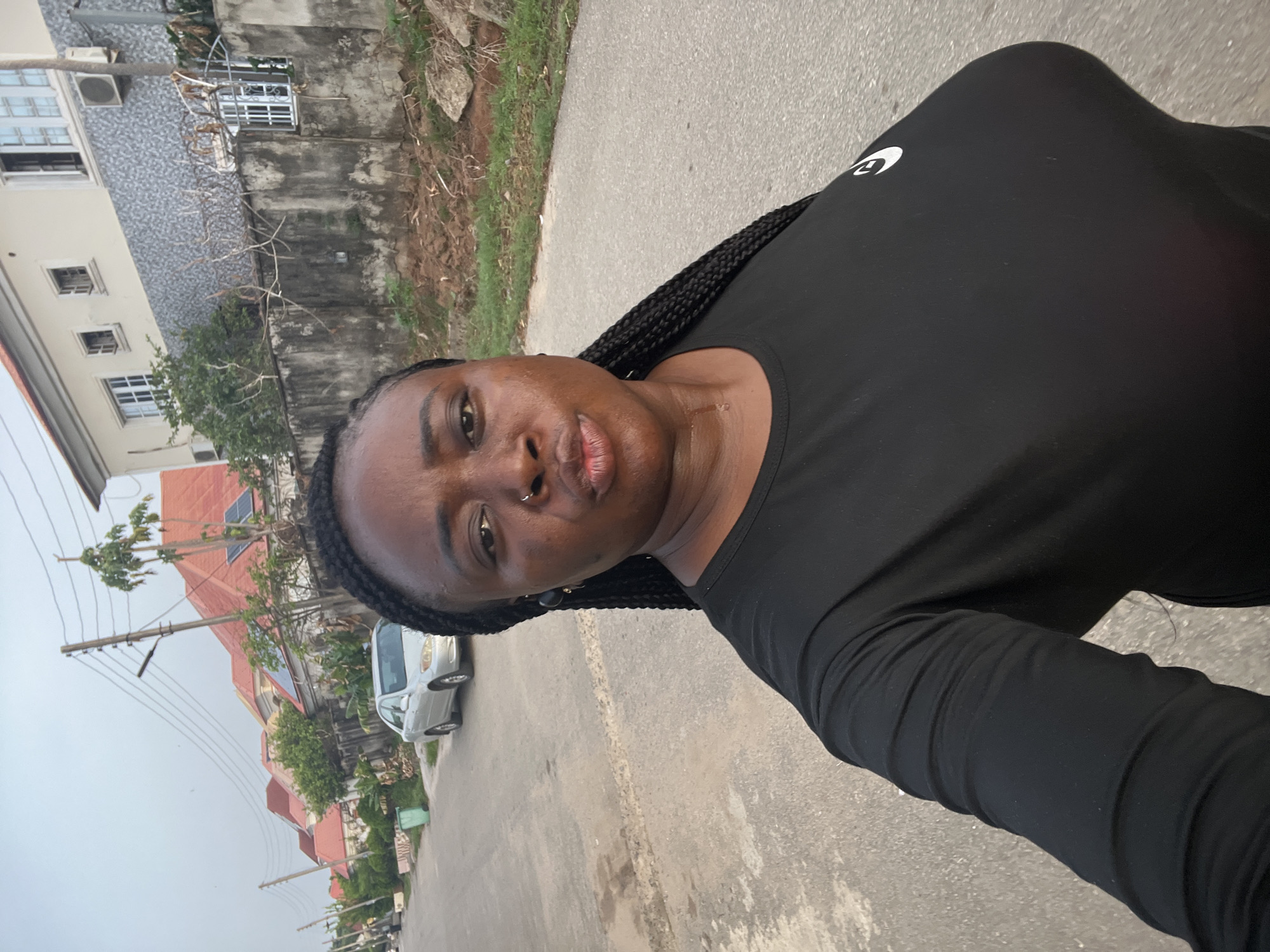 A woman with dark skin and braided hair stands on a paved road. She is wearing a black long-sleeved athletic shirt. Behind her are buildings, power poles, and a light-colored vehicle. The location is a street in what is likely a residential neighborhood of Abuja, Nigeria.