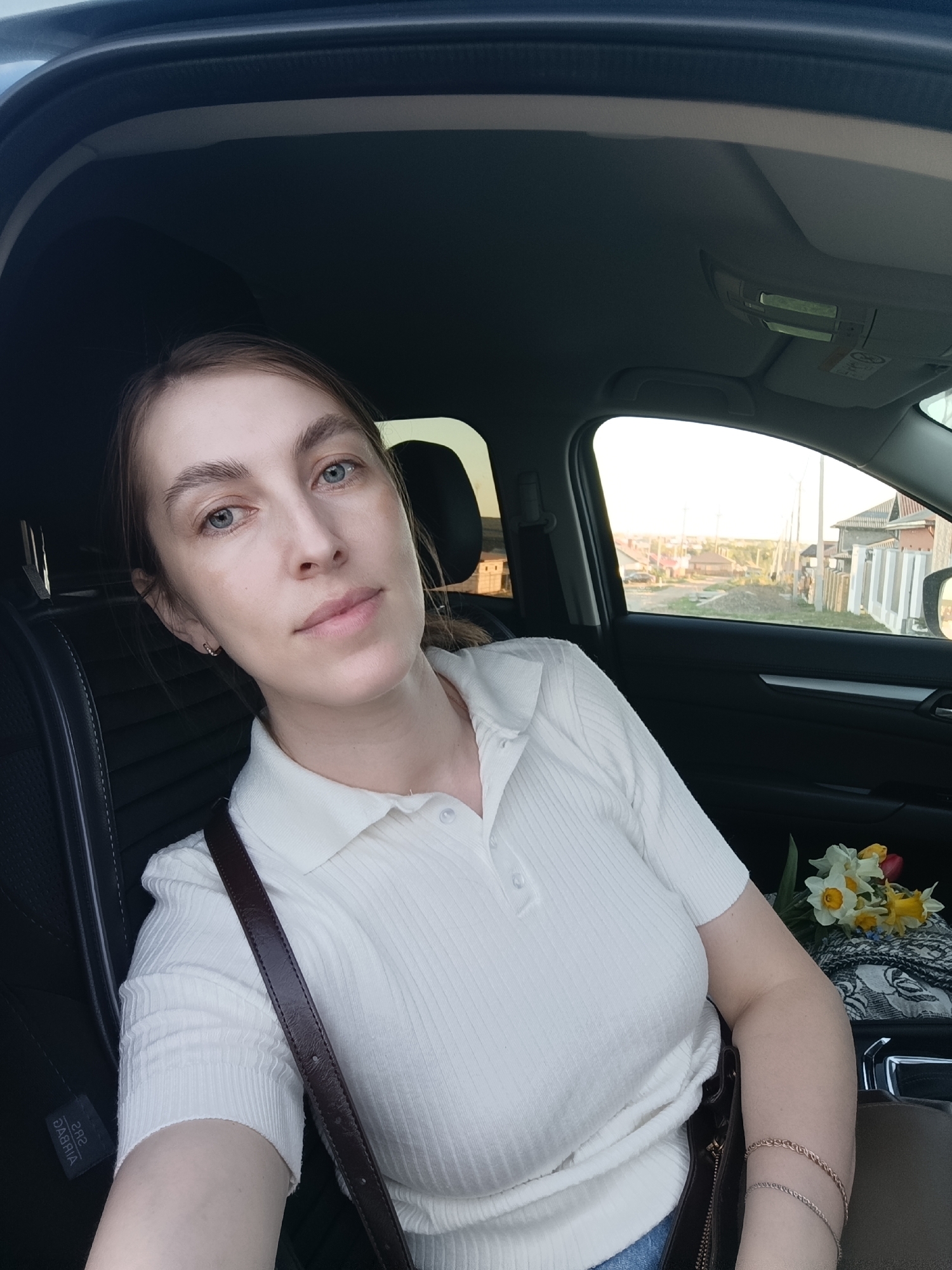 A woman is sitting in the driver's seat of a car. She is wearing a white collared shirt and has a brown purse strap across her chest. In the passenger seat, there is a bouquet of yellow and white daffodils, along with a patterned cloth. The car appears to be parked on a street in a residential area. In the background, there are houses with fences and some exposed earth. The location is likely in the city of Chaikovsky, Russia.