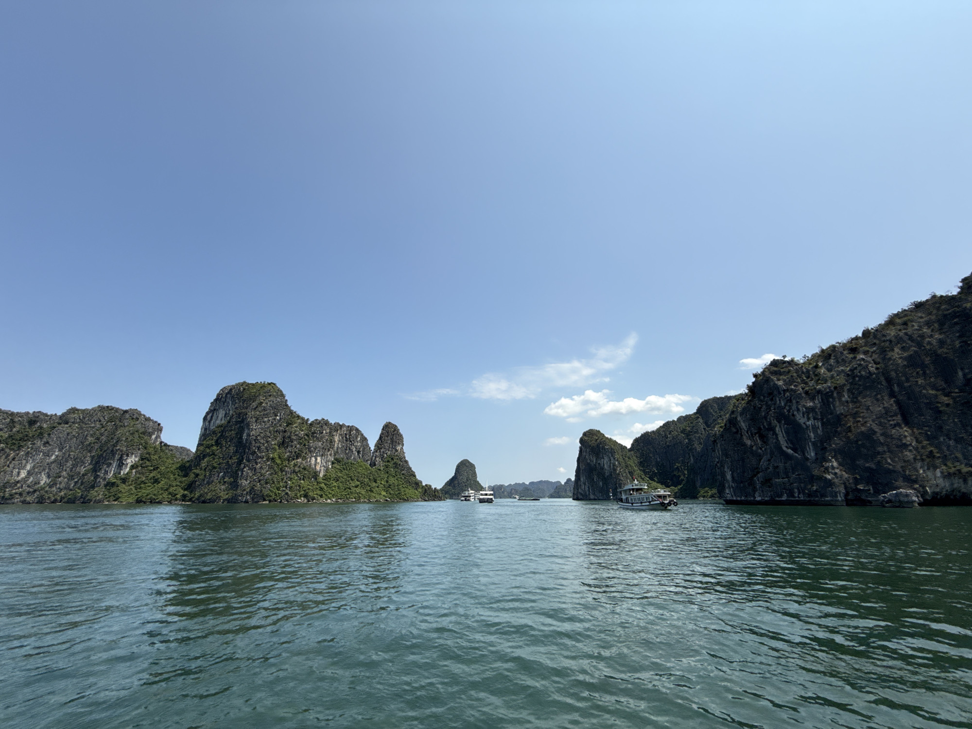 The image depicts a seascape in Tuan Chau Ward, Vietnam, under a bright blue sky with scattered white clouds. The water is dark green, showing ripples across the surface. In the distance, numerous limestone karsts jut out of the water, covered in green vegetation. Several tour boats are visible on the water, some closer to the viewer and others further away between the karsts. The overall scene suggests a popular tourist destination.