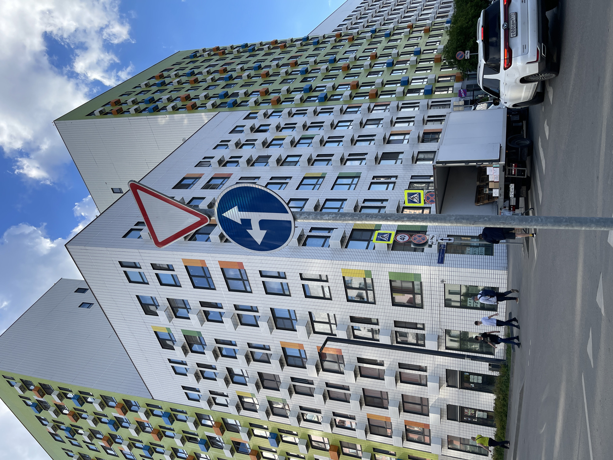 The image presents a modern urban street scene in Kommunarka, Russia, bathed in bright daylight under a blue sky dotted with white clouds. The primary subject is a tall, multi-story residential building featuring a distinctive facade. The building's lower and middle sections are covered in white or light grey tiles, while the upper section boasts a vibrant green finish. The numerous windows are framed by colorful accents in orange, blue, yellow, and green, with some appearing as small projecting elements.
In the foreground, a cluster of road signs is mounted on a pole. These include a red-bordered white triangular "Yield" sign, a blue circular sign indicating a mandatory left turn, a yellow square sign for a pedestrian crossing, and a circular speed limit sign displaying "40" km/h. Below these, a rectangular sign shows partially legible text including "ПРОЕЗД" (Passage) and numbers like "д.1-3 д.5-7", while another sign beneath it states "Коттеджный пос. Лазурный" (Cottage settlement Lazurny) and "Магазин 232" (Shop 232).
On the street, a white SUV with Russian license plate E886BM97 is parked. Behind it, a white delivery truck, likely for Ozon (with an "OZON" sign visible on the building facade near the truck), has its rear door open, revealing stacked boxes on pallets, suggesting a delivery in progress. The truck's license plate H217AB40 is also visible. Several pedestrians are walking on the sidewalk and street; three men are grouped together, one with a backpack, and another individual in a high-visibility vest is partially seen. The scene depicts a lively, contemporary residential area.