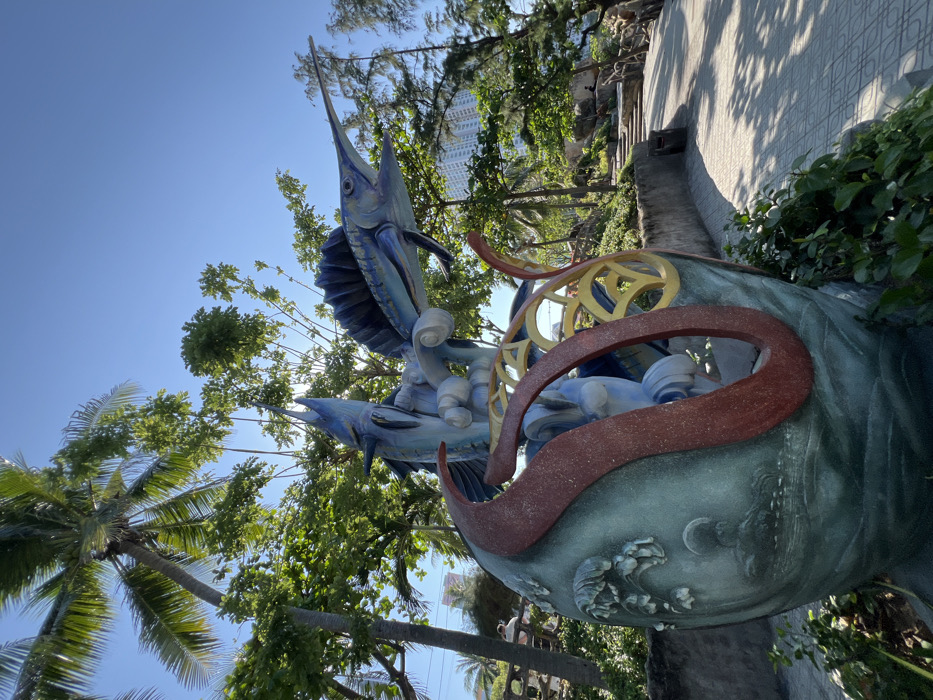This image depicts a brightly lit outdoor scene in Nha Trang, Vietnam, featuring a prominent, multi-colored marine-themed sculpture. The central subject is a large statue composed primarily of two blue marlin or sailfish, depicted leaping upwards from stylized white and blue waves. The fish exhibit pointed snouts and prominent dorsal fins. This entire marine sculpture emerges from a large, teal-green egg-shaped base, which is adorned with sculpted relief patterns depicting a seascape, including trees, water, and what appears to be a rising sun or moon motif on its lower portion. Red and yellow curvilinear, abstract elements are integrated into the sculpture, curving around the egg-shaped base.
The sculpture is situated on a paved pathway with a geometric tile pattern, leading into the mid-ground on the right side of the frame. Lush tropical vegetation, including tall palm trees and various green leafy trees, surrounds the sculpture and lines the pathway, casting shadows onto the ground. In the background, under a clear blue sky, several high-rise buildings are visible among the distant trees, indicating an urban or resort environment. To the right of the pathway, beyond a low wall, an elevated area with concrete steps is visible, leading up a tree-covered incline. A dark trash receptacle is present near the pathway on the right. In the far background, two indistinct human figures can be observed; one near a light-colored structure to the left of the path, and another further right, near the steps, with their actions not clearly discernible. The scene is illuminated by strong sunlight, suggesting daytime conditions.