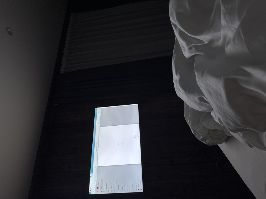 The image depicts the interior of a dimly lit room, likely a bedroom or hotel room. The specific location within Kastelruth - Castelrotto, Italy, cannot be determined from the visual evidence.
In the foreground, the lower right section of the image is dominated by white bedding, including a rumpled duvet and visible pillows, suggesting a bed. The bedding appears clean and consists of white fabric.
The central subject is a large, illuminated rectangular display or projection on the far wall. This display shows a web browser interface with "youtube.com" in the address bar. The screen is actively loading, indicated by a circular loading animation and the text "Loading your videos" centered on the white page. A white mouse pointer, shaped as an arrow, is visible near the bottom center of the displayed page. The browser also shows the YouTube logo and navigation elements on the left sidebar. The wall behind the display is dark, consistent with vertical wood paneling or a similar dark textured surface.
To the right of the displayed content, in the midground and background, a sheer, light-colored curtain is visible, drawn across what appears to be a window or alcove. The curtain is translucent, allowing minimal light penetration, and hangs from the ceiling. The overall lighting in the room is low, with the screen being the dominant light source. A single recessed downlight fixture is visible on the dark ceiling in the upper right quadrant of the image. No people are visible. No active interactions or specific actions are depicted beyond the screen's loading state.