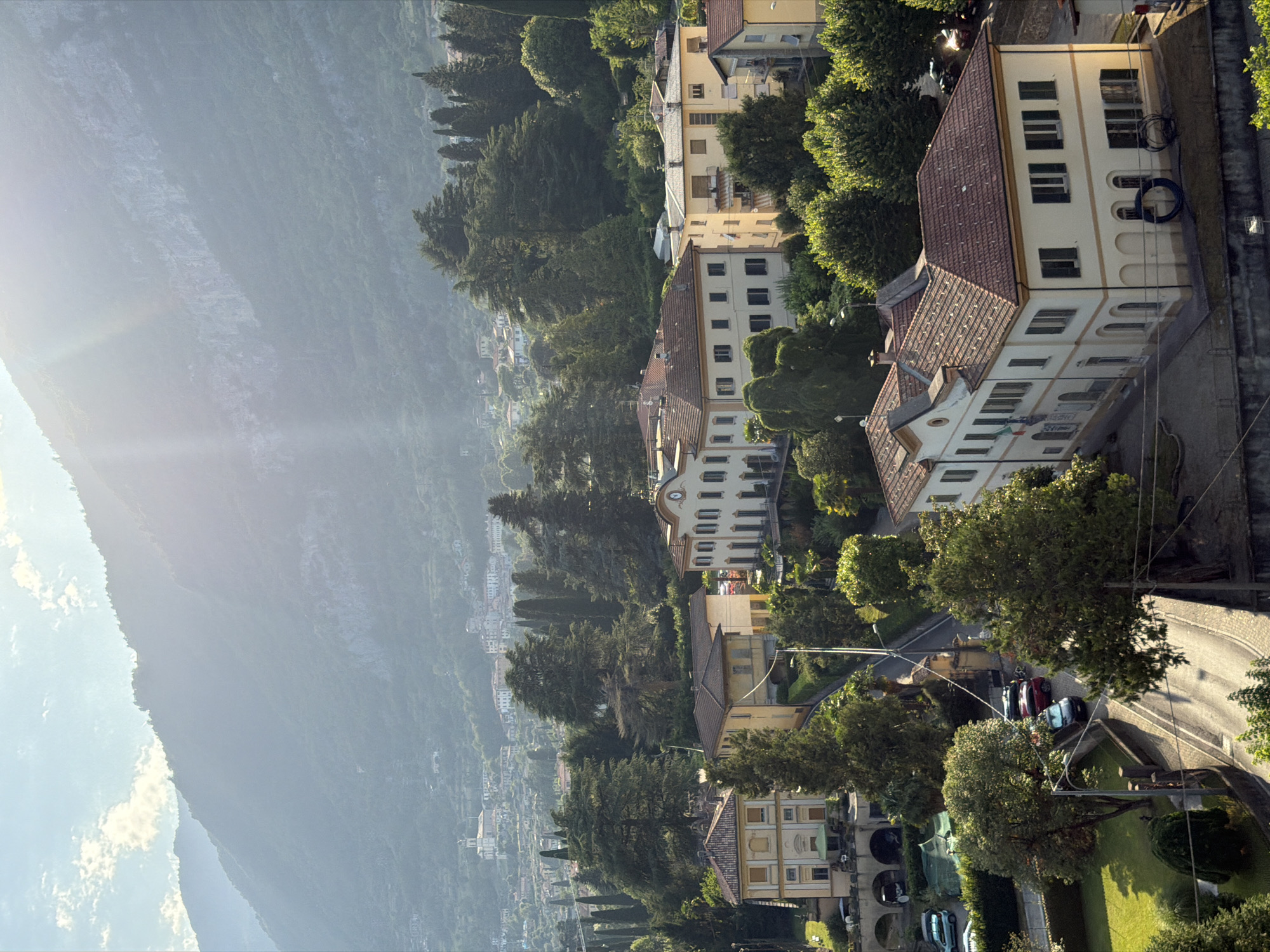 The image captures a sun-drenched view of Tremezzina, Italy, with a focus on residential buildings nestled amongst lush greenery and a mountainous backdrop. In the foreground, a cream-colored building with a red tiled roof and green shutters is prominent, featuring multiple arched windows on the ground floor. Adjacent to it, another building, also with a red tiled roof, exhibits a yellow facade and dark shutters. To the left, a pale yellow building with decorative trim and dark shutters is visible. A winding road leads through the scene, with several cars parked along its edges. Beyond these structures, a sprawling town with white buildings and red roofs extends up the hillside towards the imposing, tree-covered mountains. The sky is bright blue with scattered clouds, and sunlight streams in from the upper right, casting lens flare across the image. The overall impression is one of a serene, European hillside community bathed in warm, natural light.