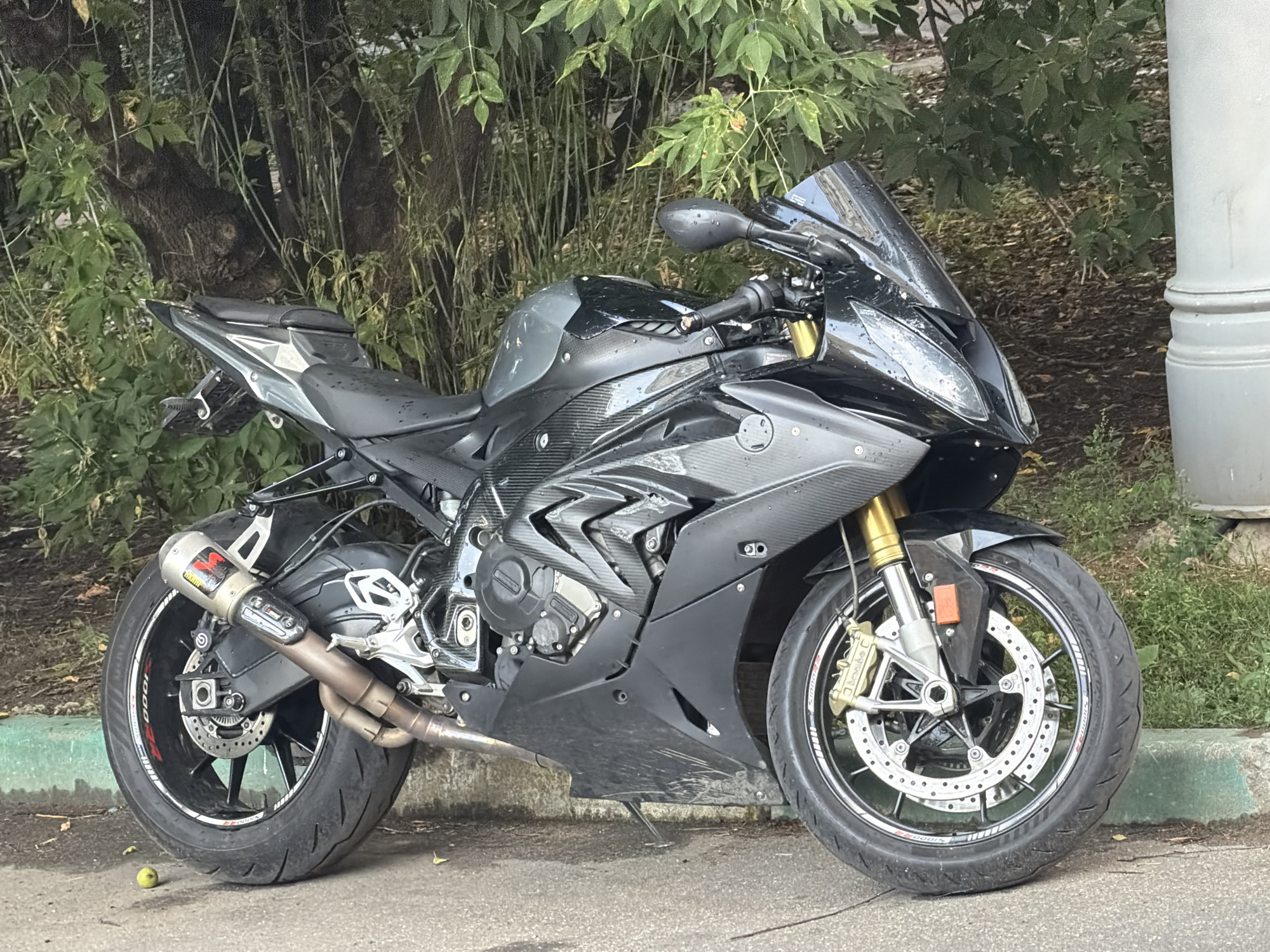 A black and grey BMW S1000RR motorcycle is parked on a paved surface next to a grassy embankment and a tree. The motorcycle is facing towards the right side of the frame. Its front wheel and part of the engine are visible, along with its exhaust pipe and rear wheel. The motorcycle appears to be in good condition, with no visible damage. The surrounding environment is lush and green, with trees and foliage creating a natural backdrop. A light-colored, cylindrical object, possibly a lamppost or pillar, is present to the right of the motorcycle. The scene suggests an outdoor setting, likely in a park or along a roadside in the city of Khimki, Russia.